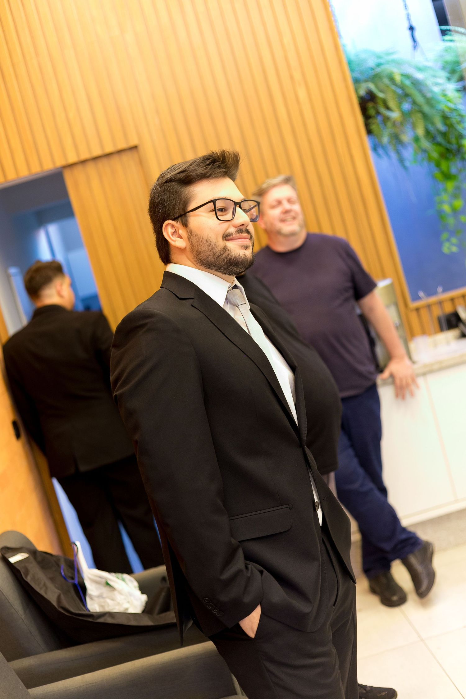 A young man in a black suit and glasses smiles confidently in a modern lobby with people in the background.