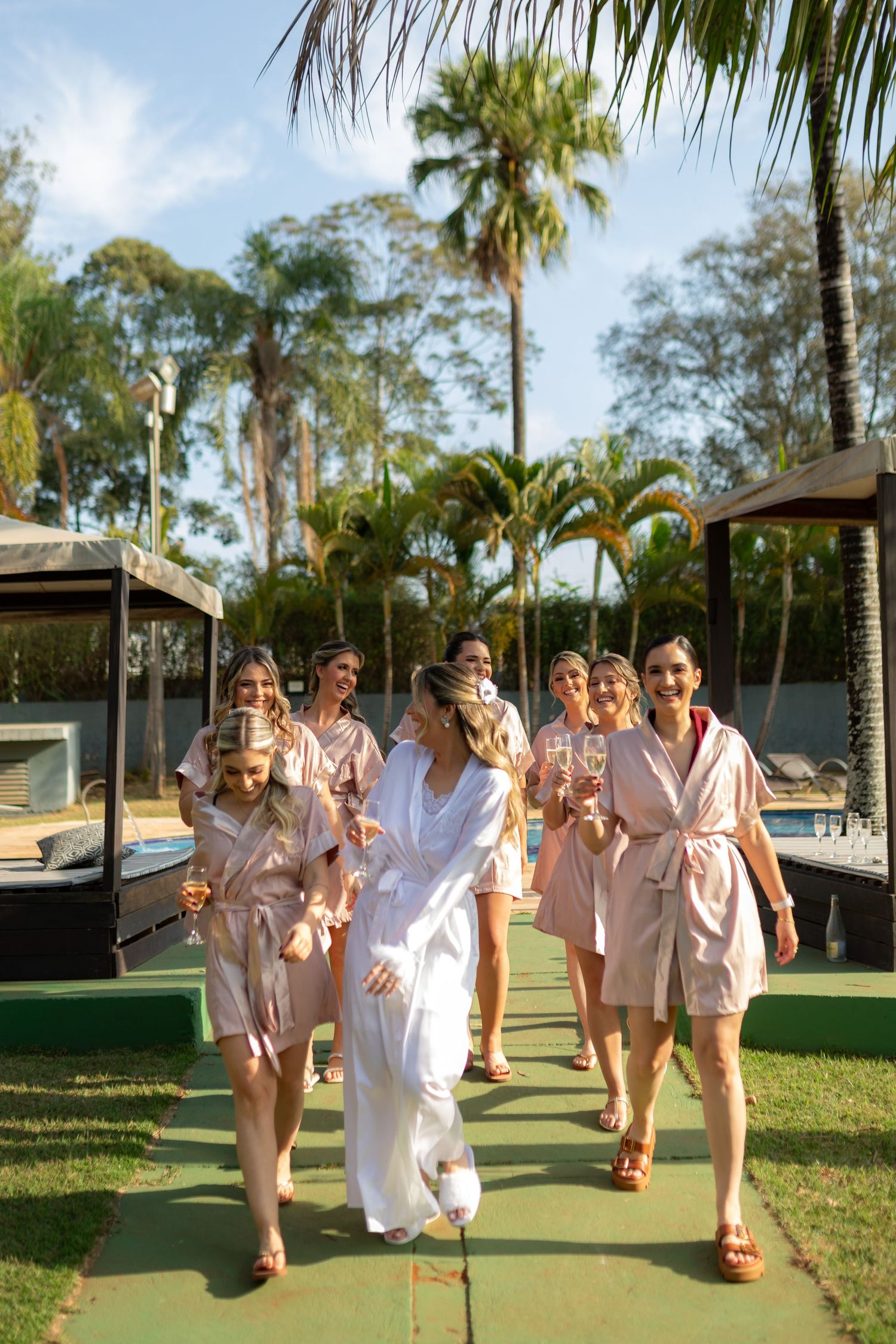 Bride in white robe walks with bridesmaids in blush robes holding champagne glasses by a tropical pool.