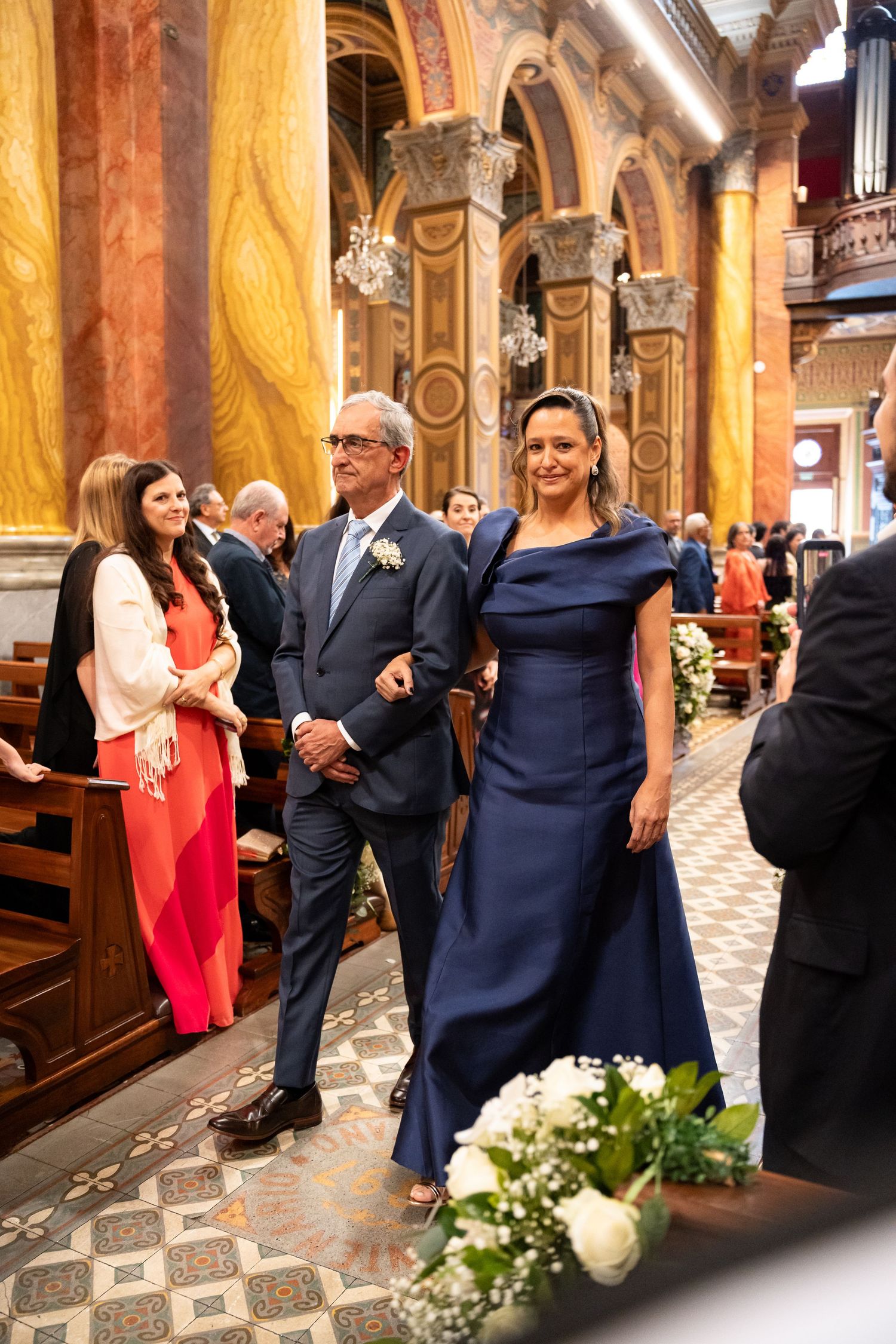 Elegant couple walking down church aisle at wedding ceremony, woman in navy blue gown, man in gray suit.