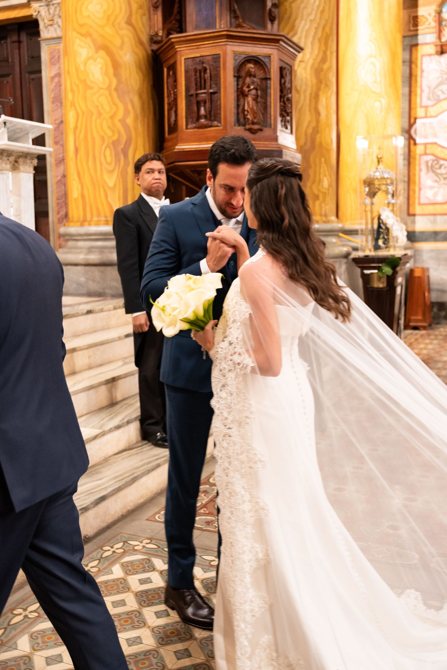 Groom in navy suit kisses bride's hand at church altar, bride wearing lace gown and flowing veil holds white roses.