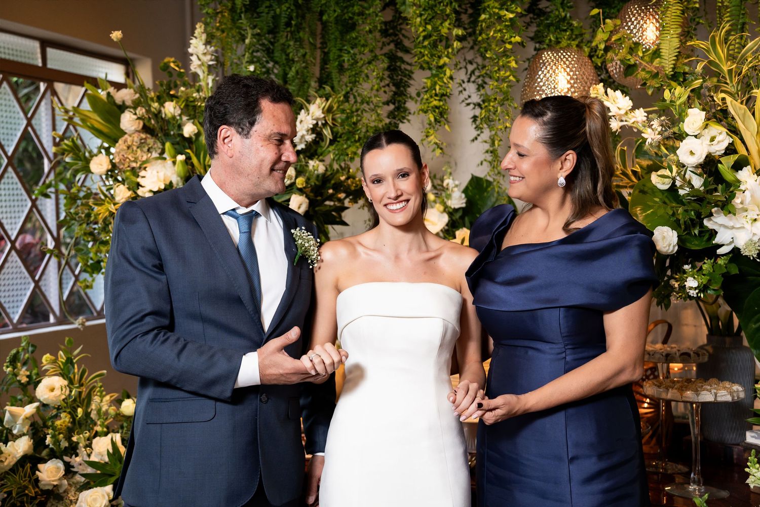 Bride in white strapless gown smiles with two guests at floral wedding ceremony backdrop.