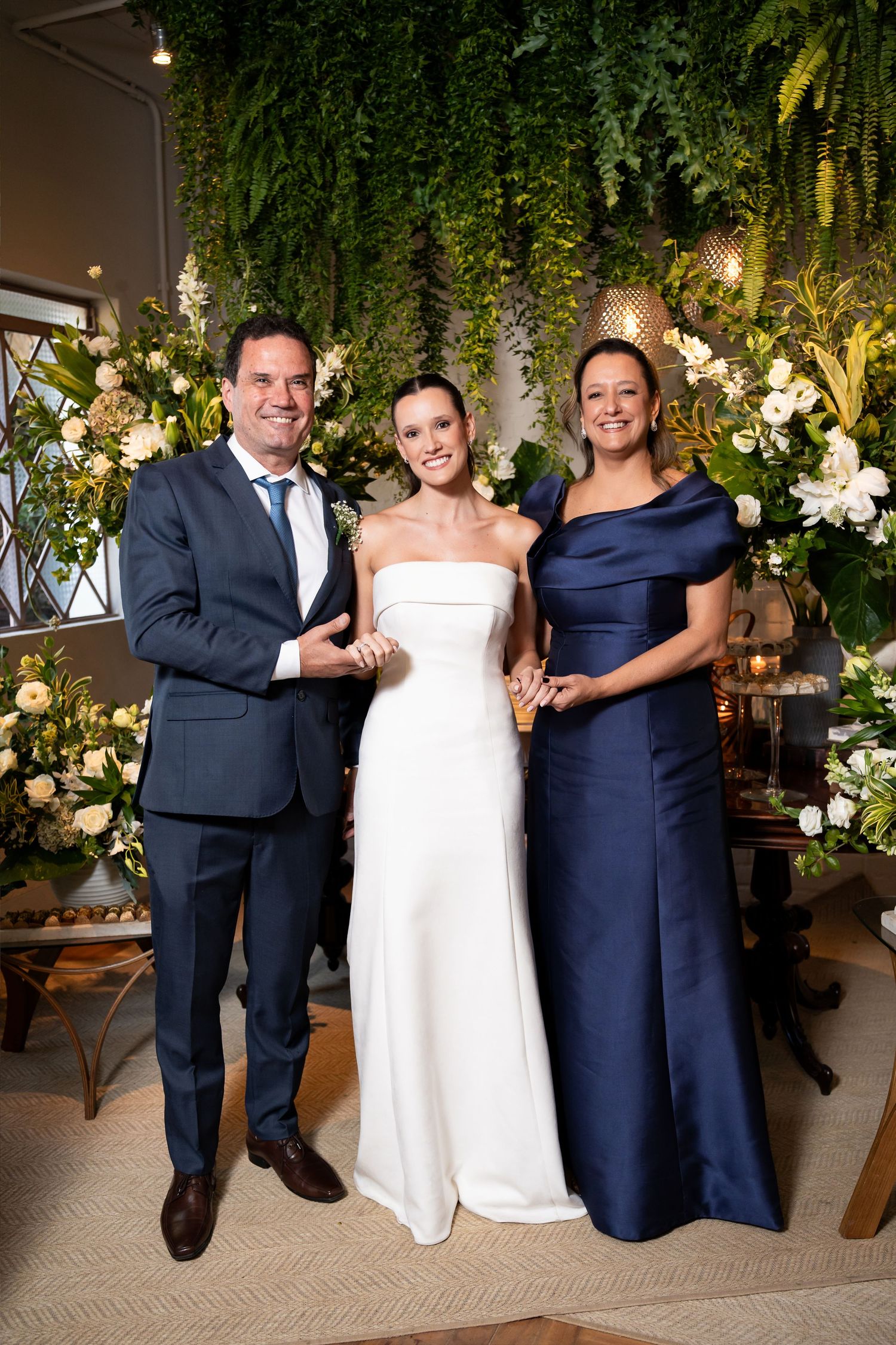 Bride in white strapless gown poses with man in navy suit and woman in navy dress before floral backdrop.