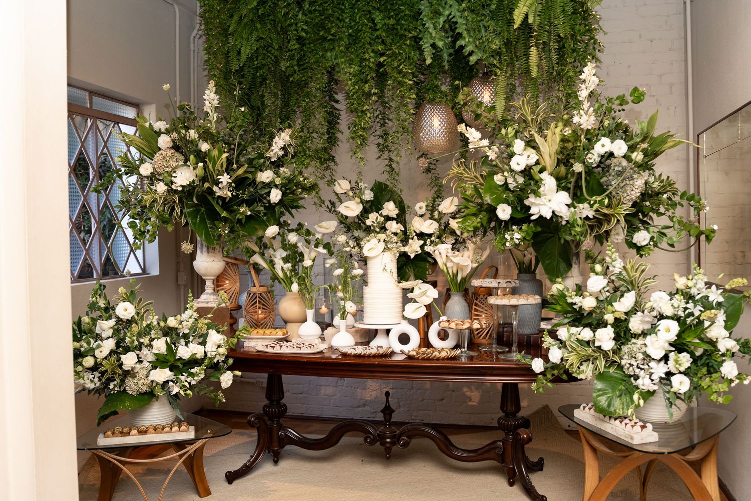 Elegant wedding dessert table adorned with lush white floral arrangements and hanging greenery in a luxurious setting.