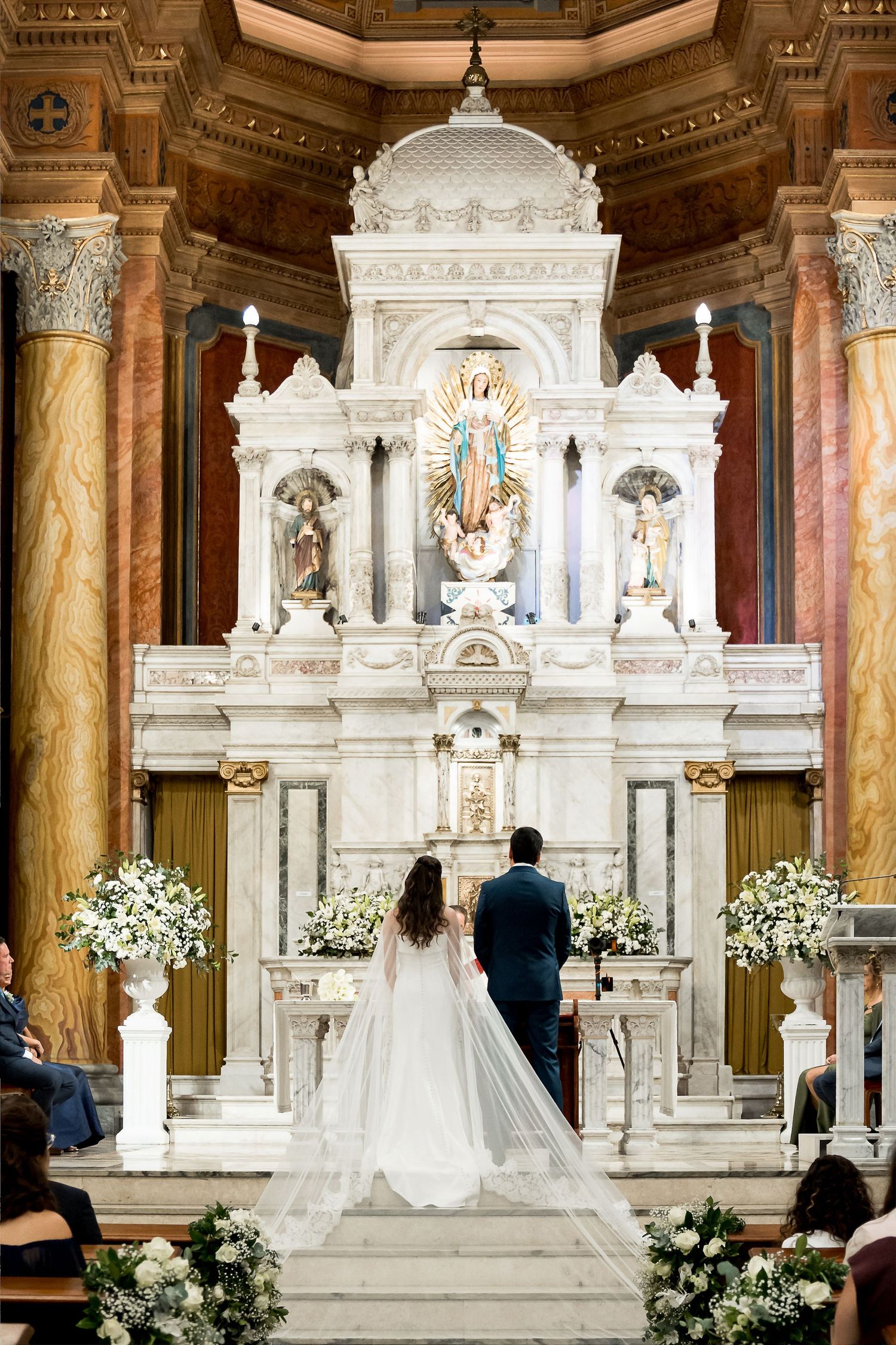 Bride and groom stand before ornate white marble altar with Virgin Mary statue in elegant church wedding ceremony.