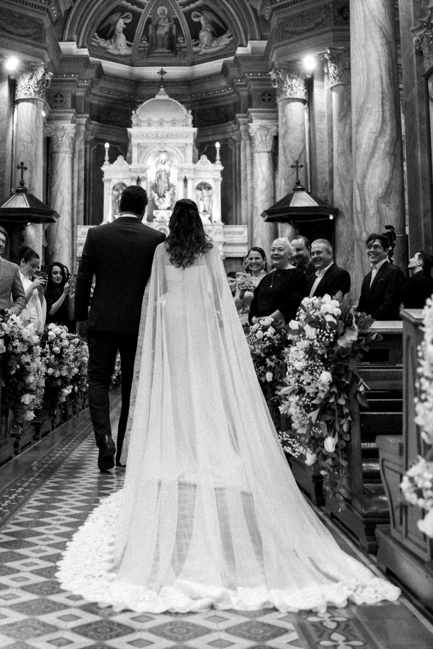 Black and white photo of bride in flowing cape gown walking down church aisle with groom during wedding ceremony.