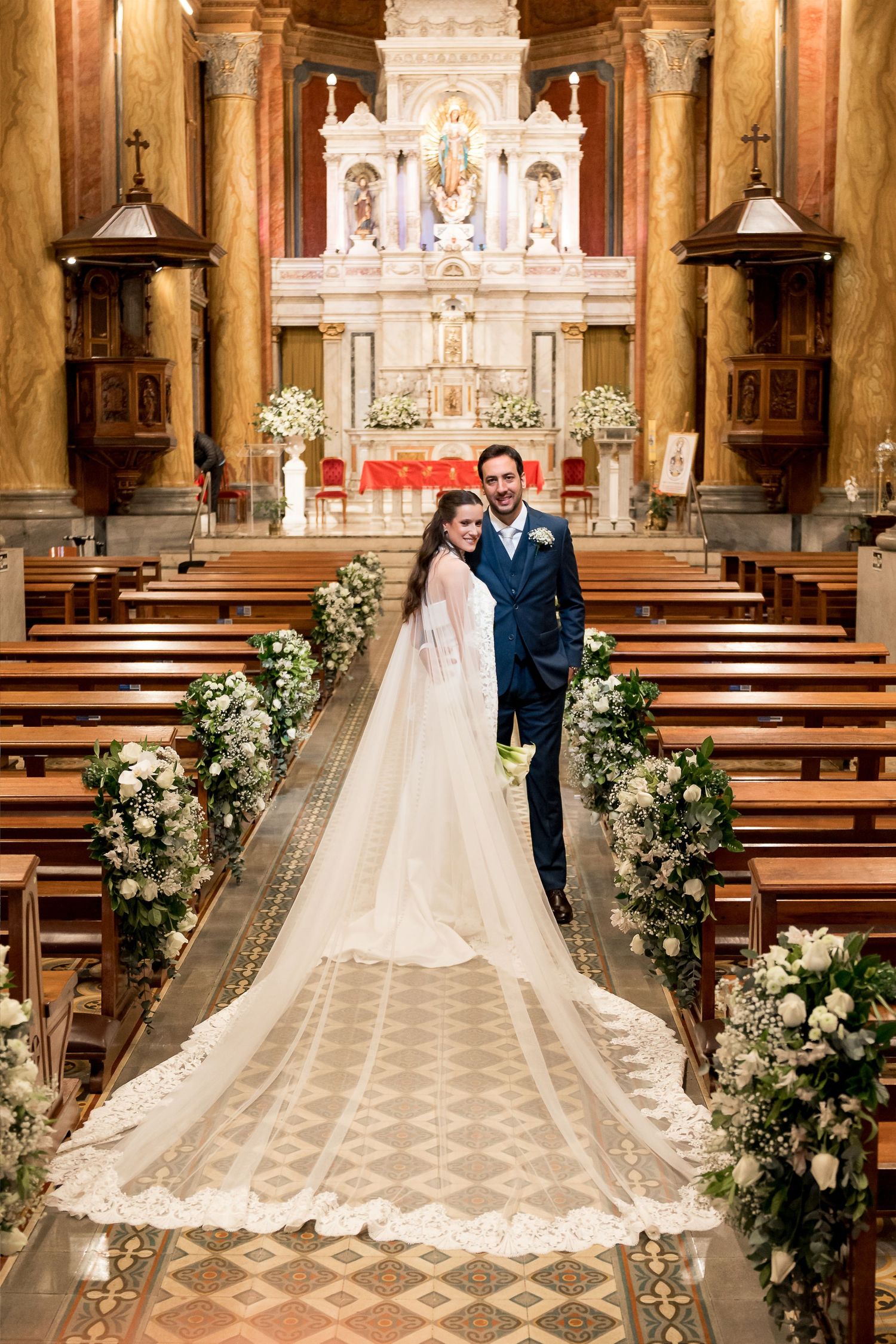Bride and groom pose at church altar after wedding ceremony, with floral arrangements lining the aisle.