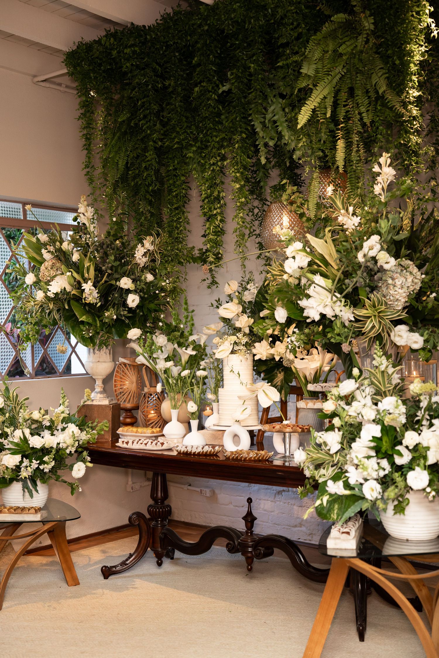 Elegant wedding dessert table adorned with white flowers, greenery, and a tiered cake beneath lush hanging ferns.