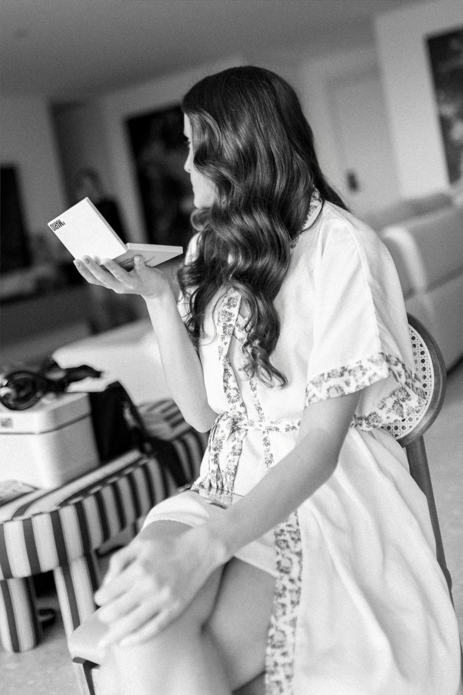 Black and white photo of a bride in a floral robe reading a card while getting ready on her wedding day.