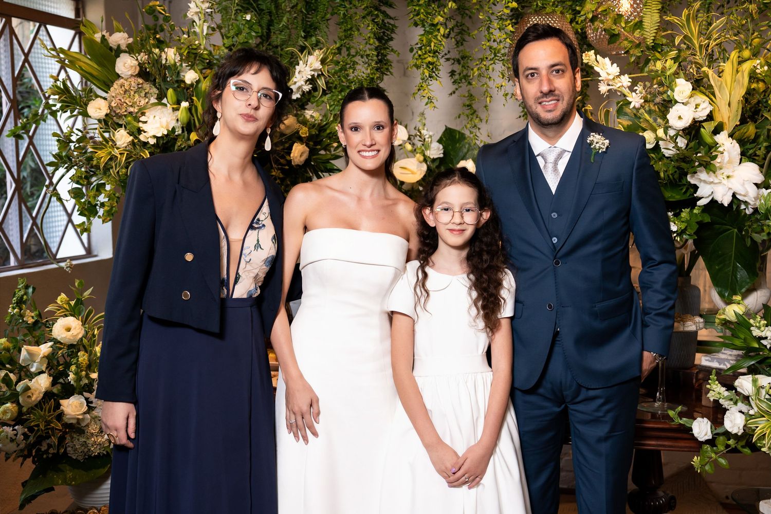 Wedding party of four posing before floral backdrop, bride in white gown with man in suit and two women.