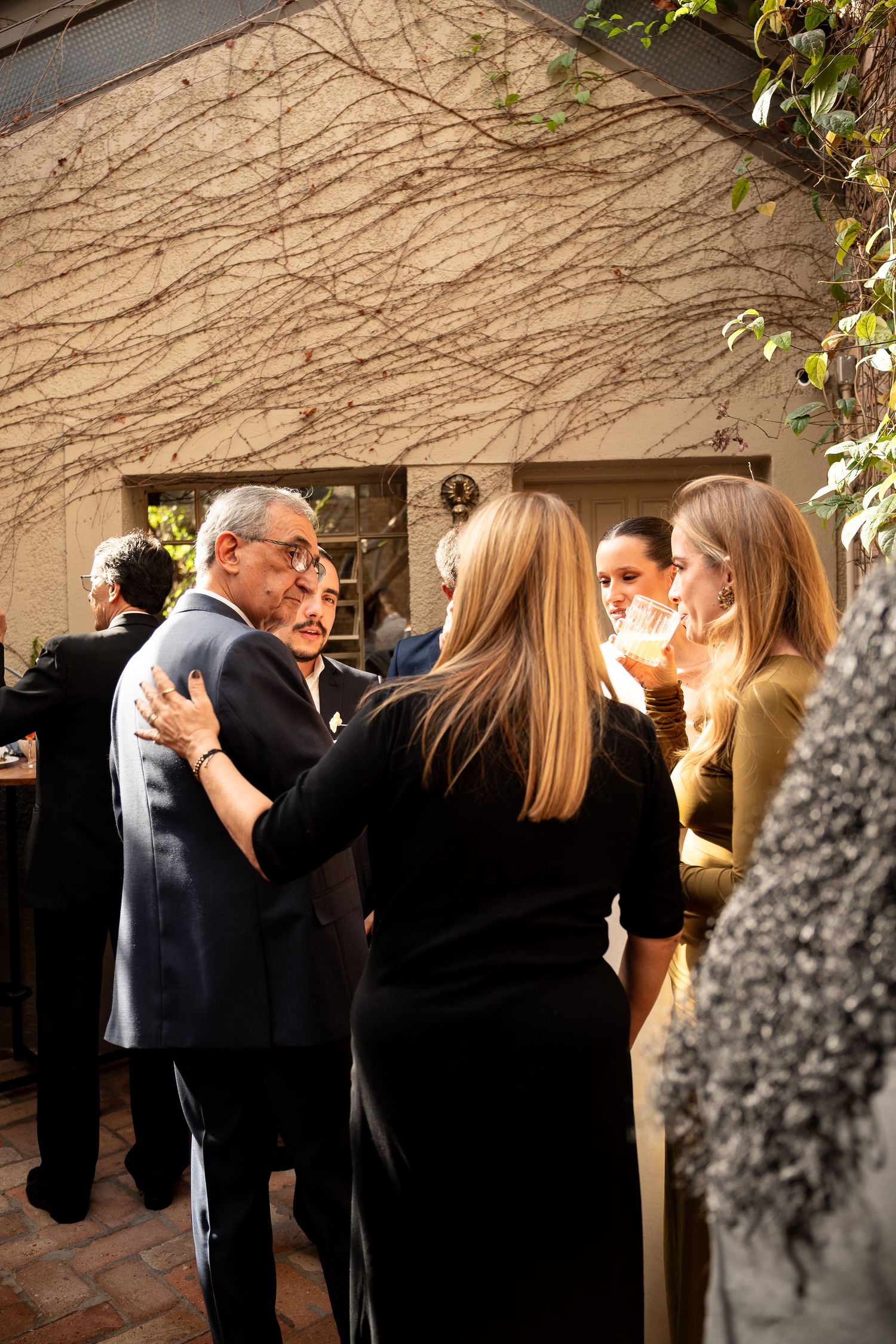 Guests mingling outdoors at an elegant evening gathering near a building with bare vine-covered walls.