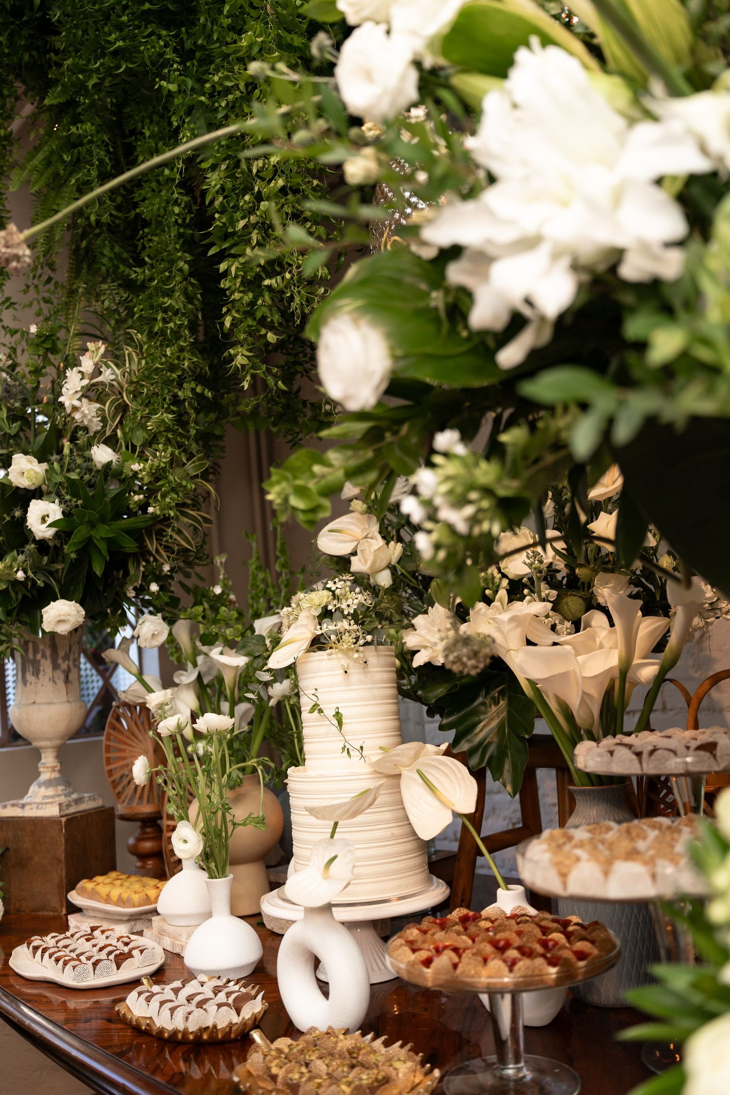 Elegant wedding dessert table with tiered white cake, floral arrangements, and assorted sweets against a lush green backdrop.