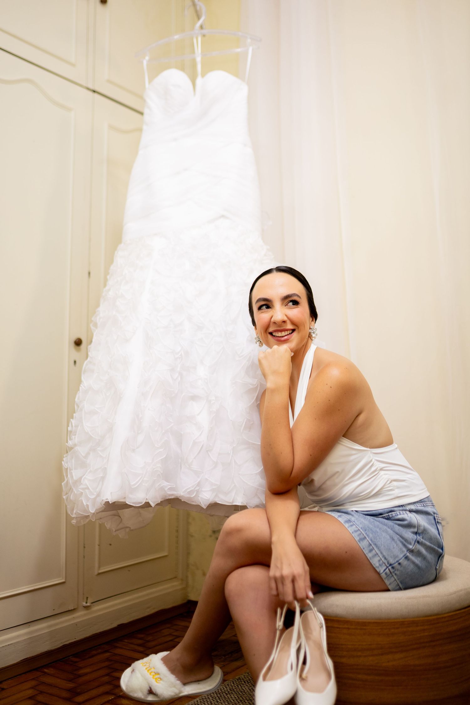 Smiling bride-to-be sitting beside her white lace wedding dress hanging on a wardrobe, holding bridal heels.