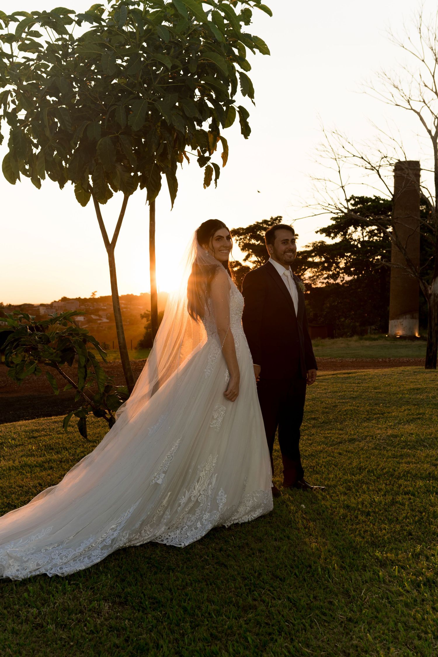 Bride in white lace gown and groom in black suit pose at golden sunset on green lawn near trees.