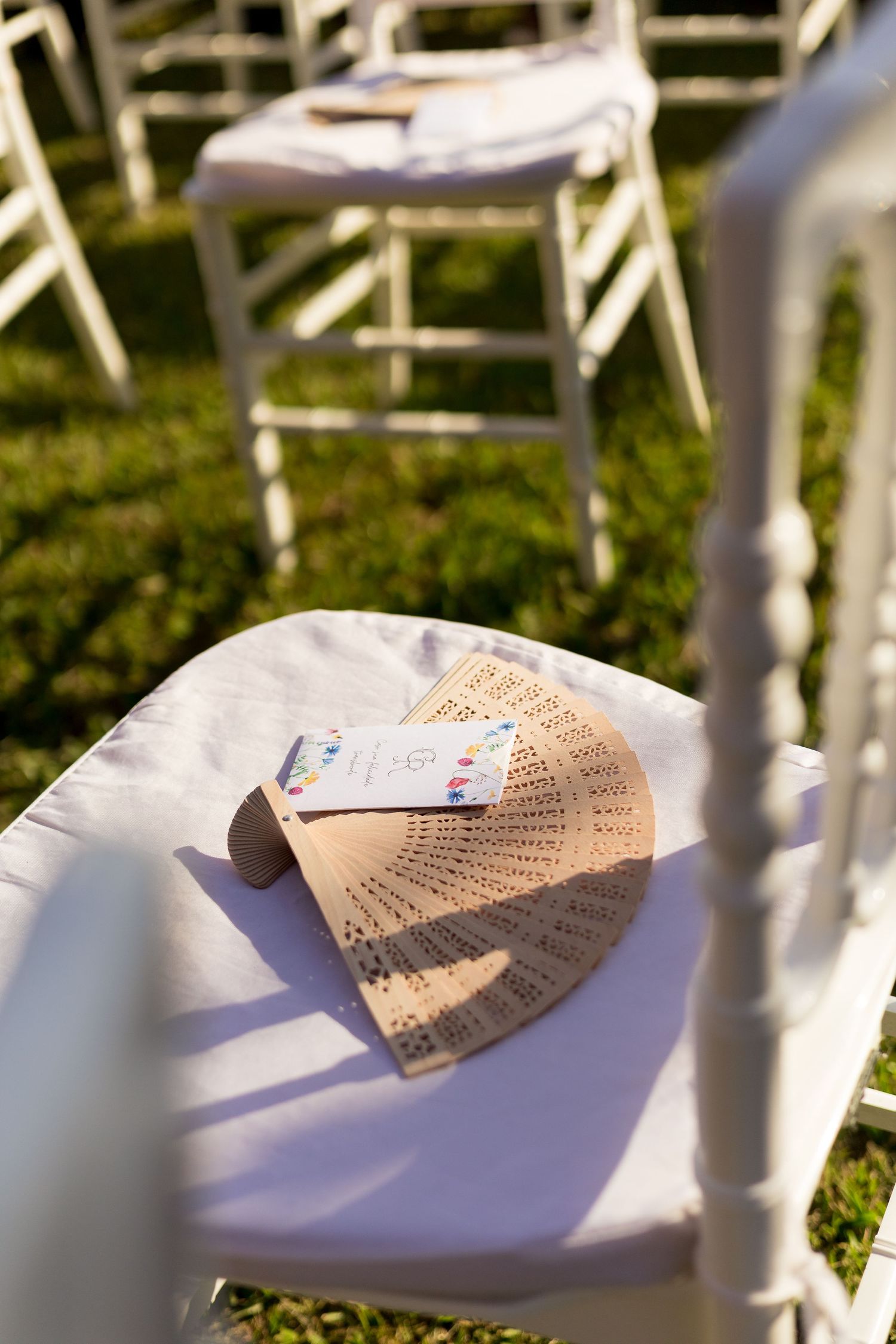 Wooden hand fan and wedding program on white chair cover at outdoor ceremony on green grass.