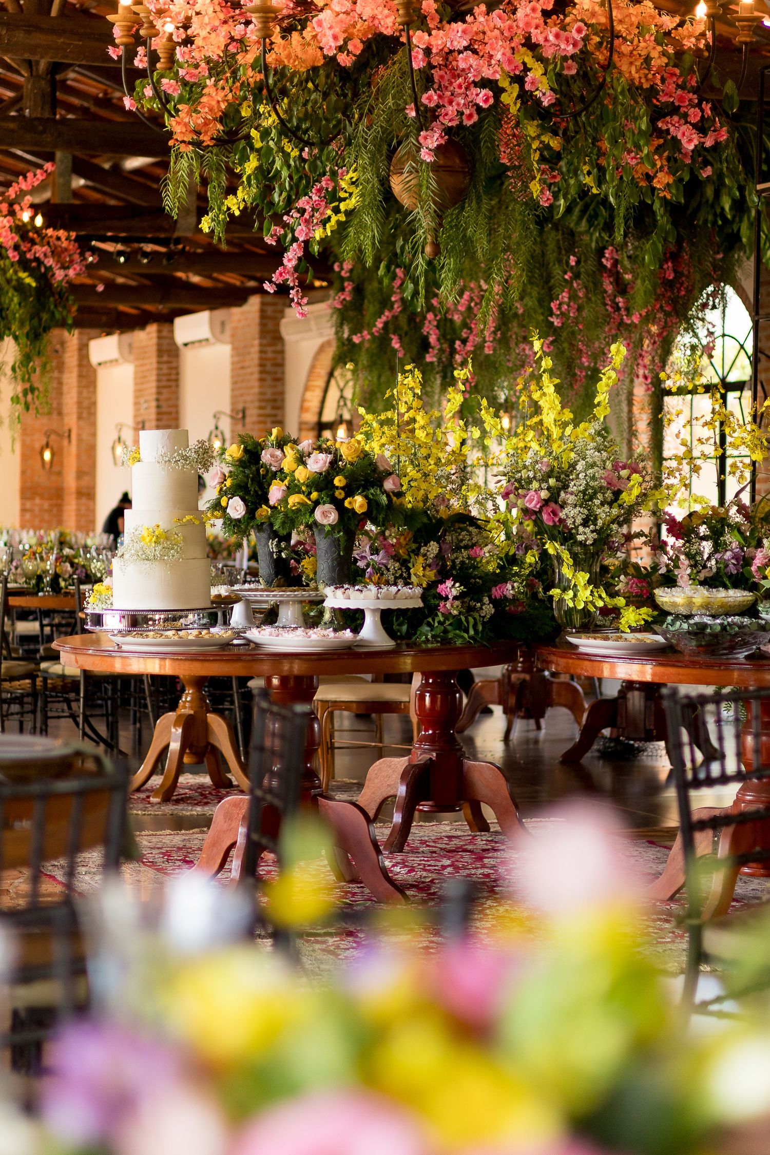 Elegant wedding reception with tiered cake on wooden table, surrounded by lush floral arrangements and hanging flowers overhead.