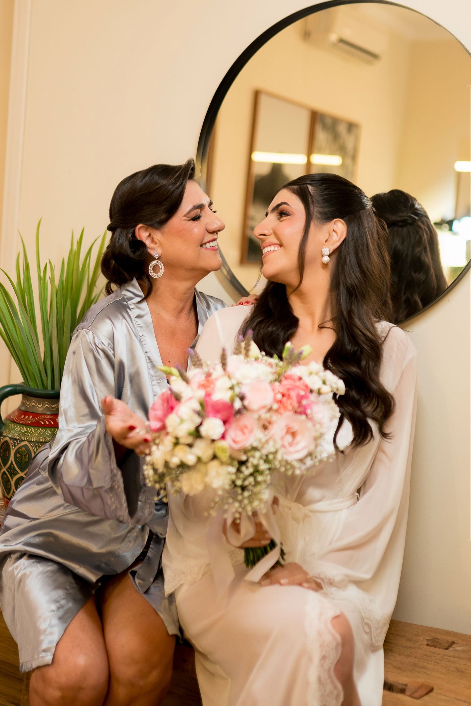 Bride and bridesmaid in robes smiling at each other, holding a pink and white floral bouquet in front of a round mirror.