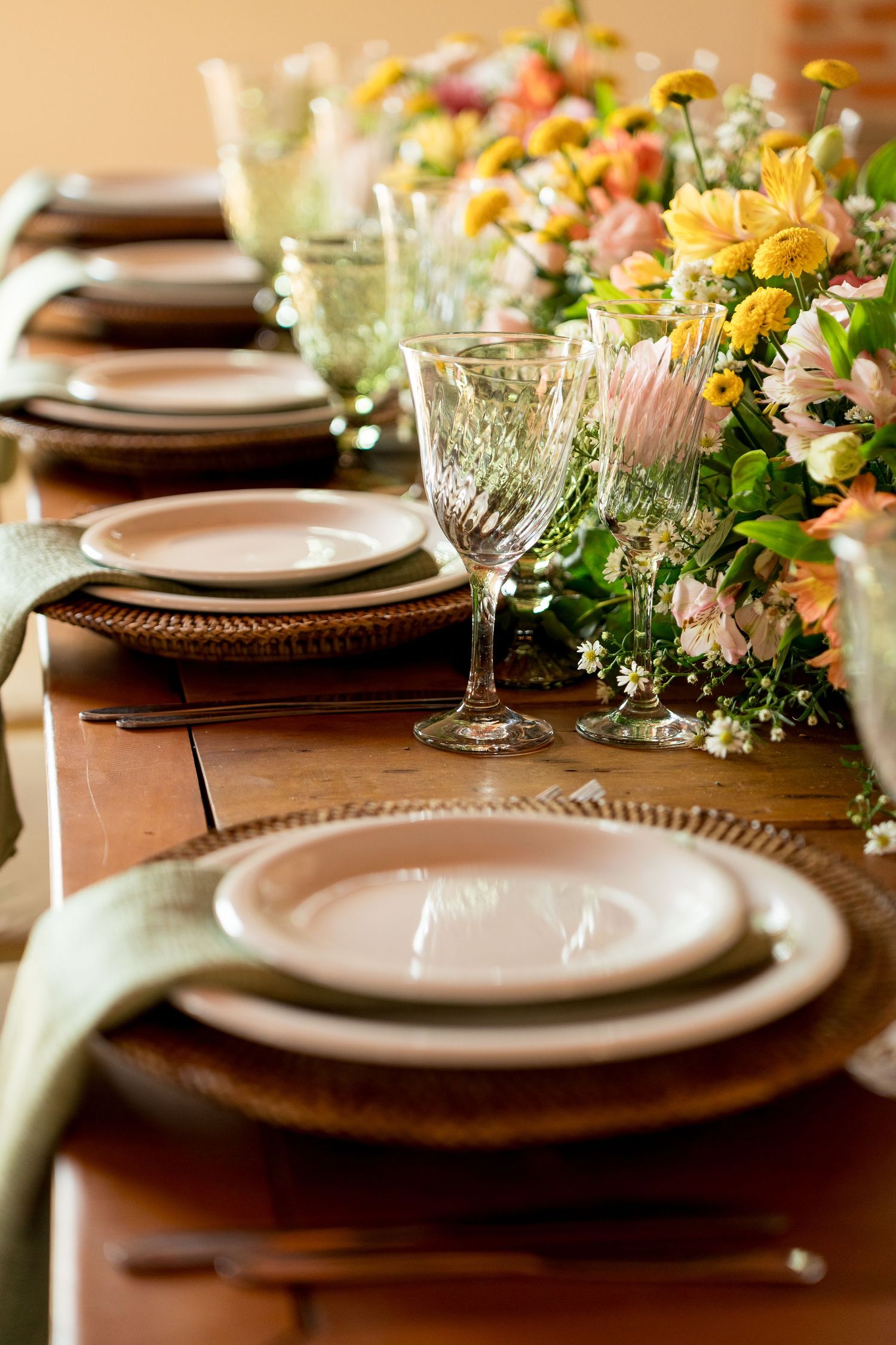 Elegant rustic wedding table setting with crystal glasses, stacked plates, and a floral centerpiece of yellow and pink blooms.