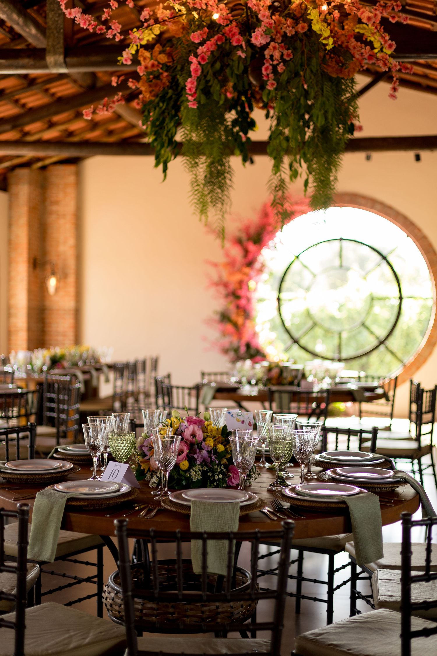 Elegant wedding reception with floral ceiling installation, round tables, chiavari chairs, and a circular window backdrop.