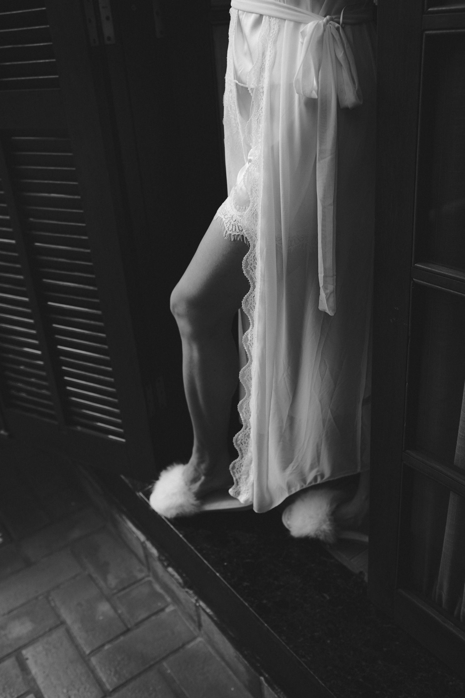 Black and white photo of a woman in a sheer white lace robe and fluffy slippers standing by shuttered doors.