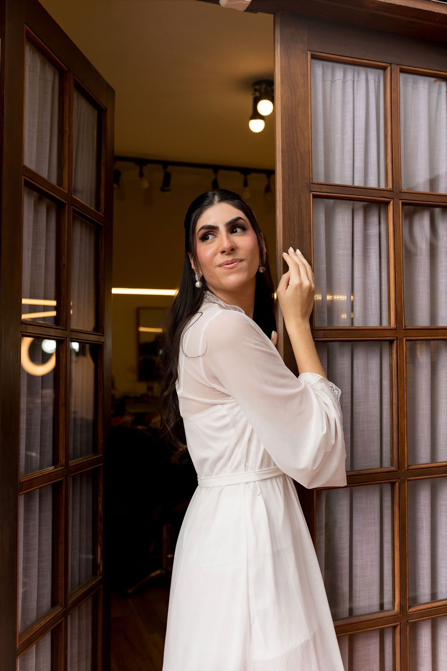 Woman in white dress standing in wooden doorway, looking over shoulder with elegant earrings and dark hair.