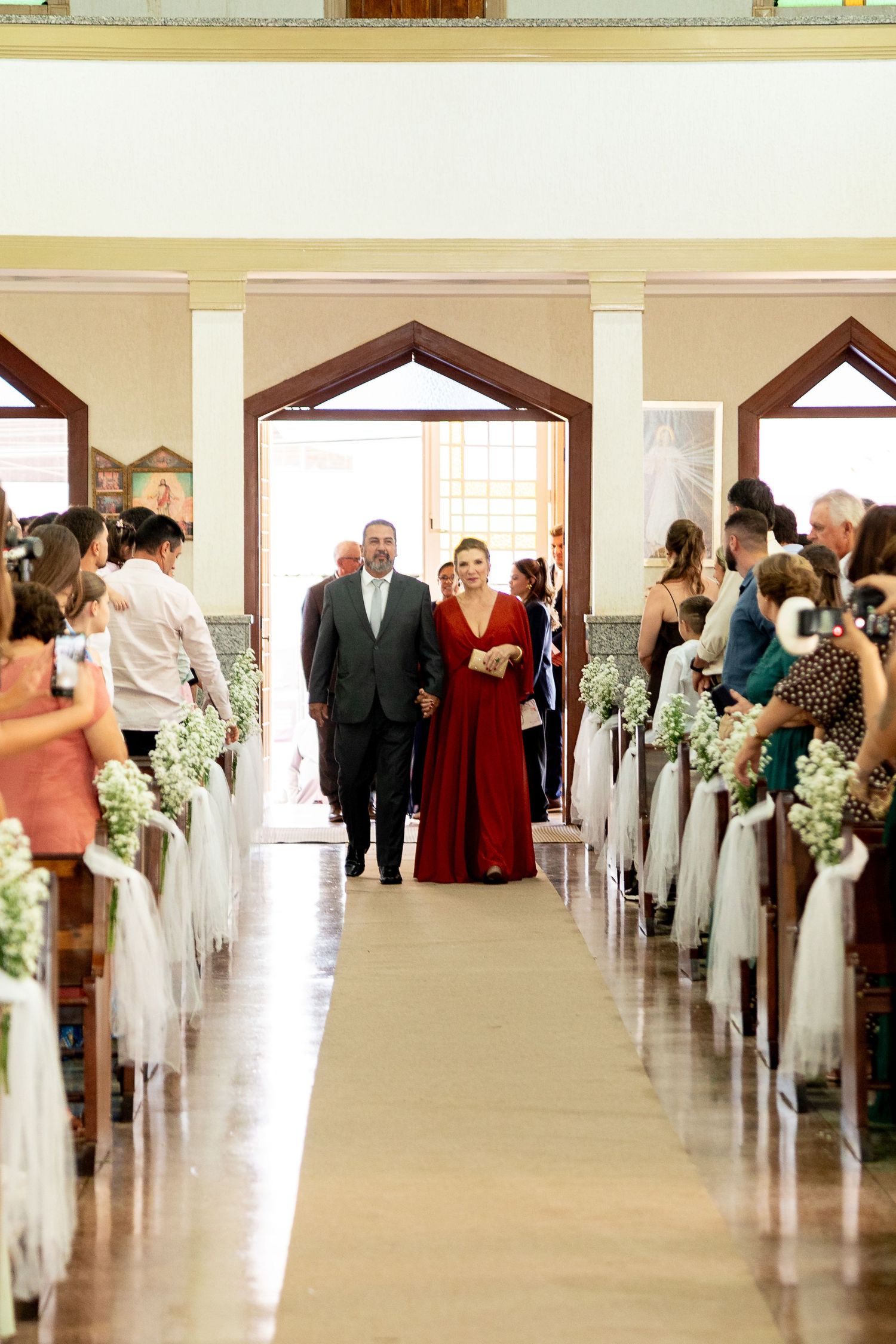 Woman in red gown walks down church aisle with escort during elegant wedding ceremony with floral pew decorations.
