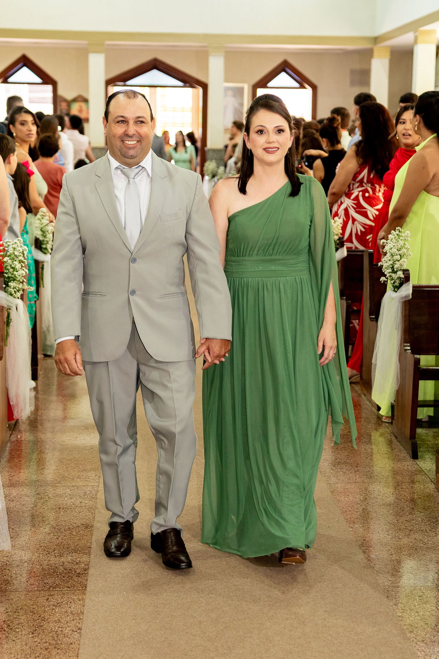 Smiling couple in gray suit and green dress walk hand in hand down wedding aisle.