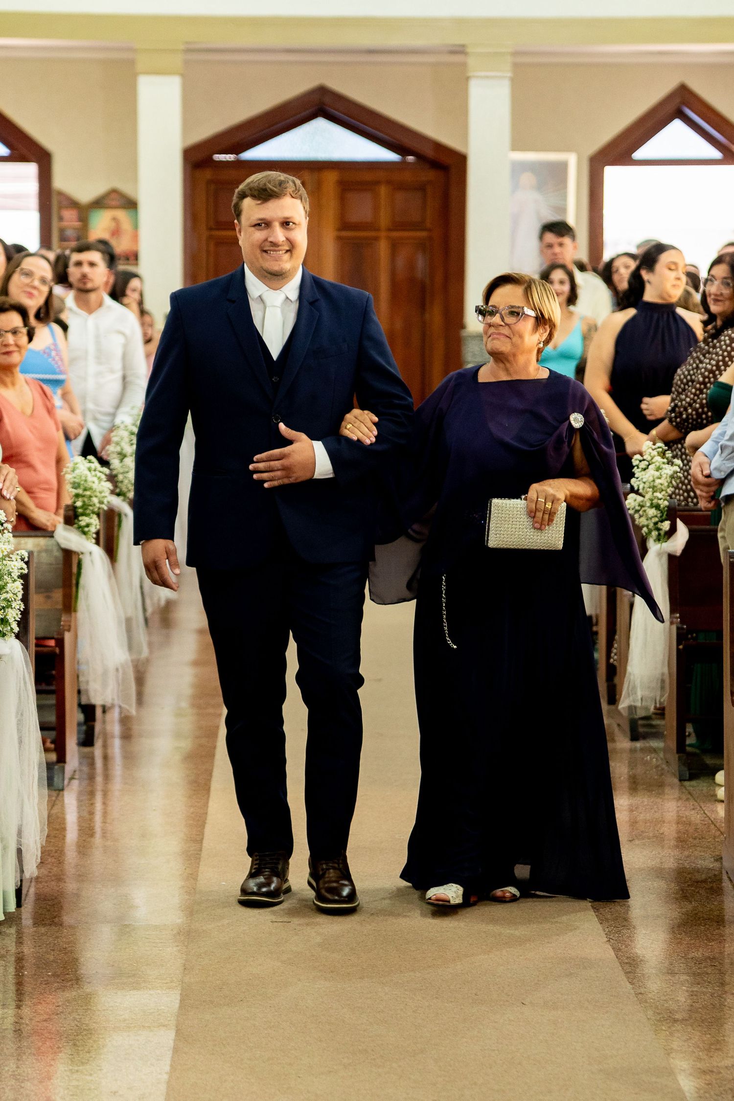 Groom in navy suit walks down church aisle with older woman in purple gown during wedding ceremony.