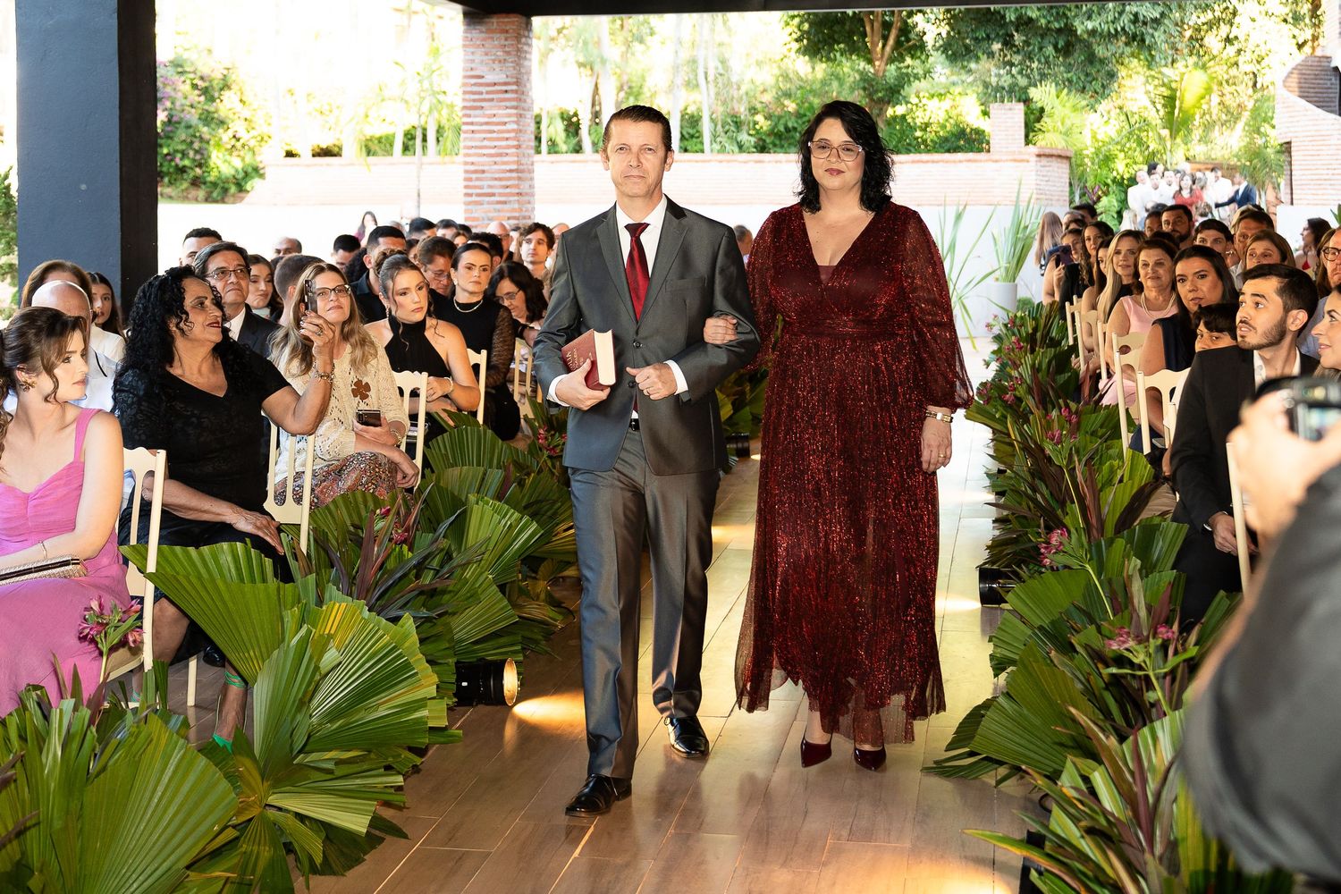 Couple walking down floral-lined wedding aisle surrounded by seated guests at an elegant outdoor ceremony.