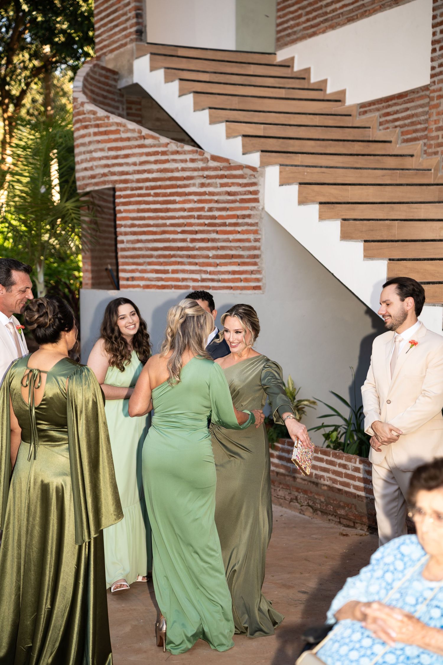 Bridesmaids in green dresses greet guests at an outdoor wedding venue with brick stairs and lush greenery.