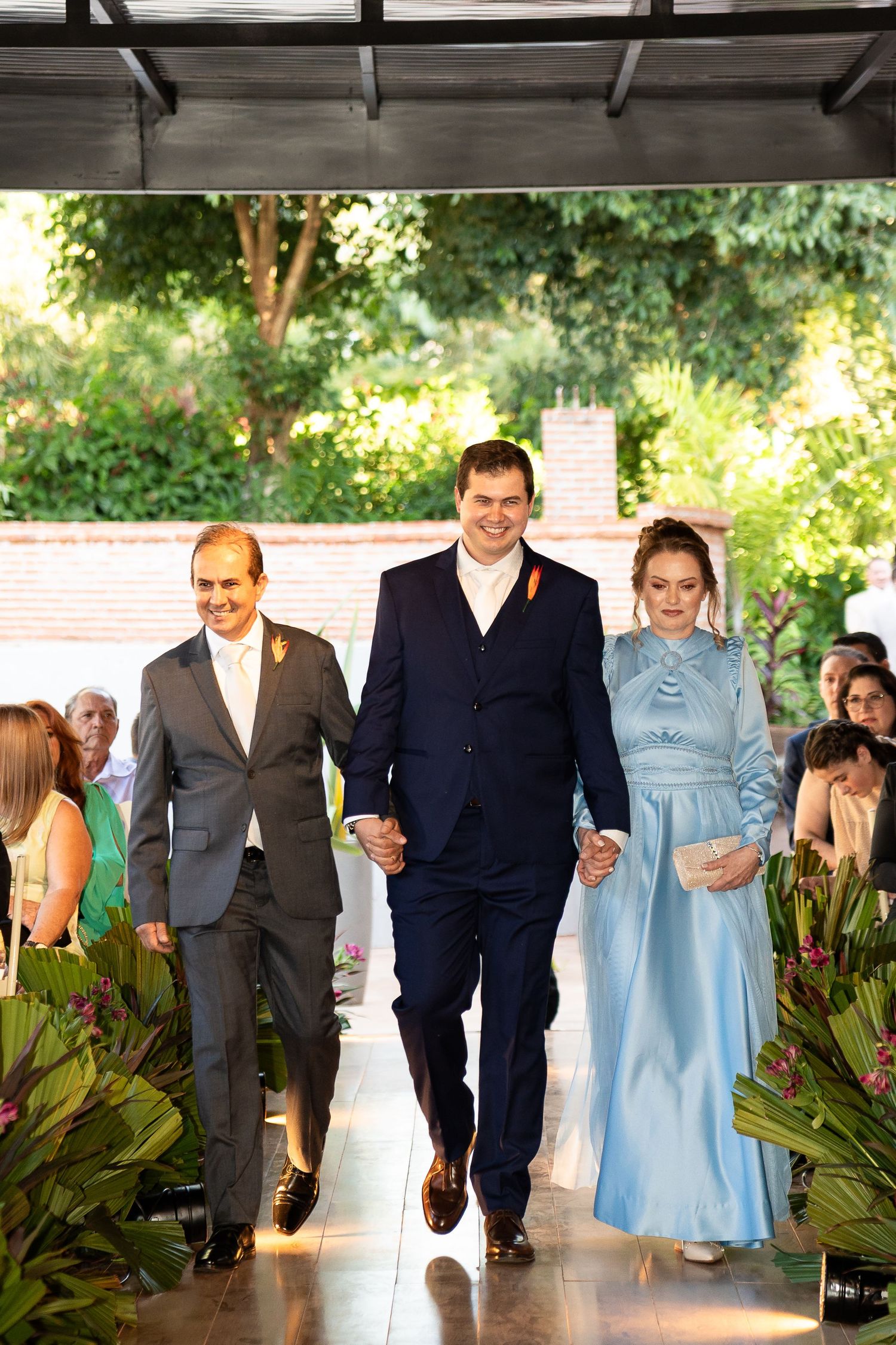 Groom in navy suit walks down aisle with parents at outdoor wedding ceremony, woman in light blue gown.