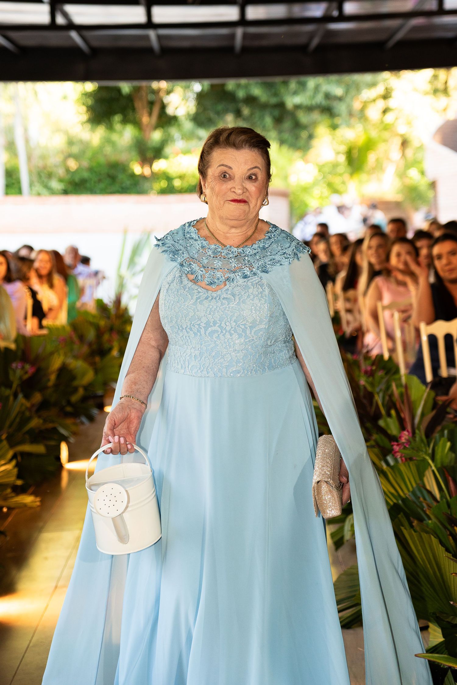 Elderly woman in elegant light blue gown with cape walks down aisle at formal outdoor event.