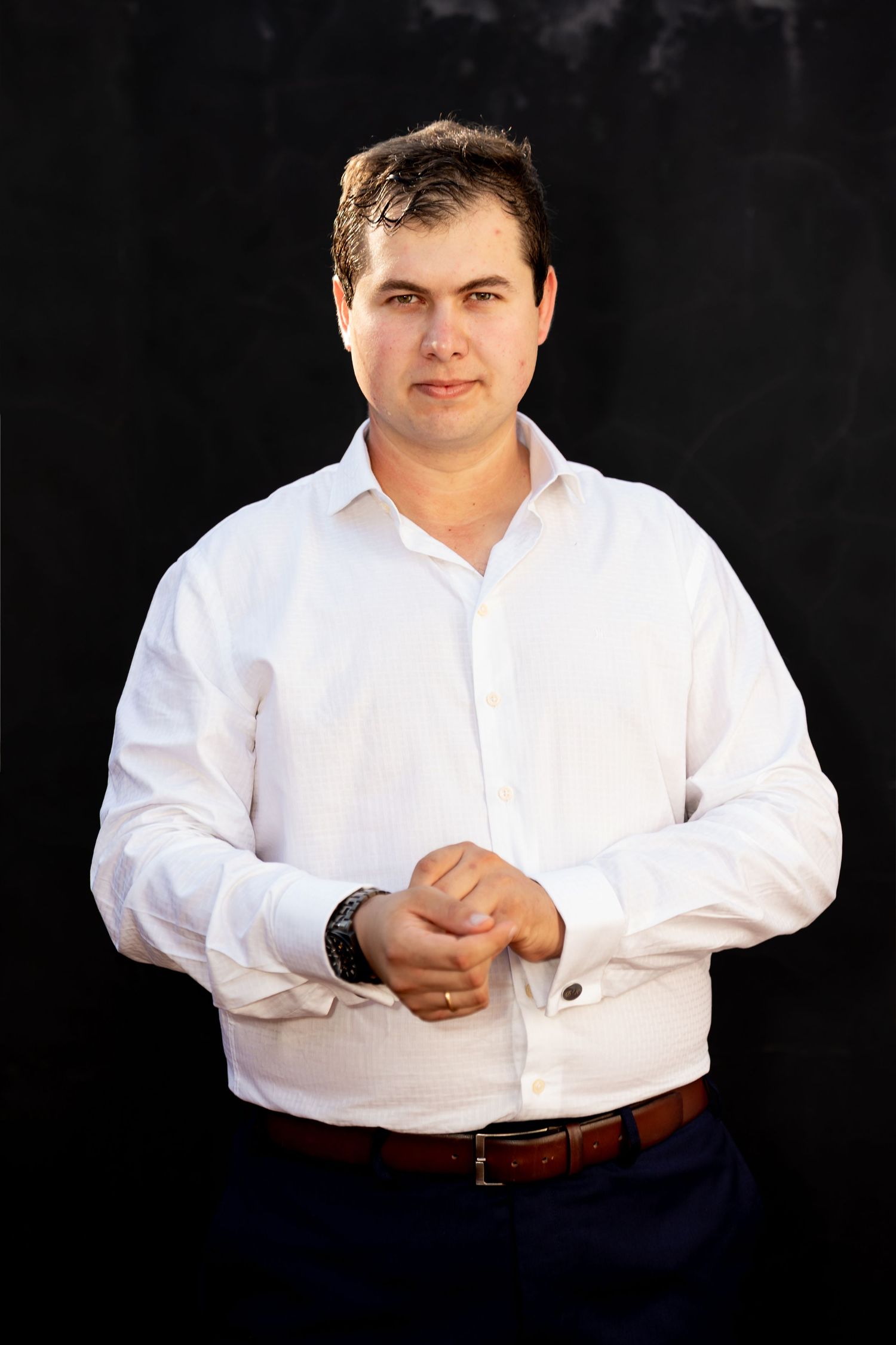 Professional man in white dress shirt and brown belt posing confidently against dark background.