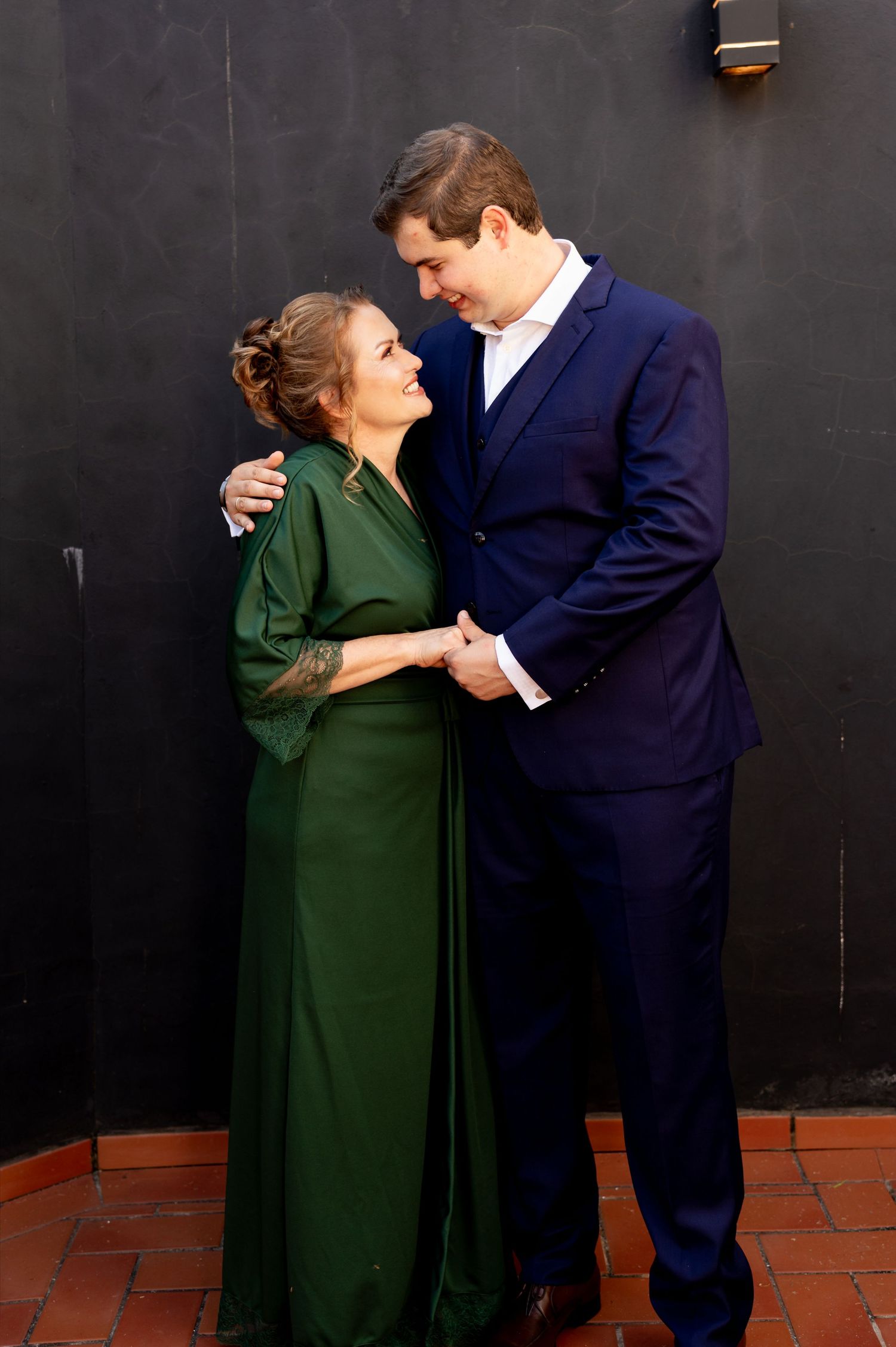 A woman in a green gown and a tall man in a navy suit share a tender moment against a dark wall.