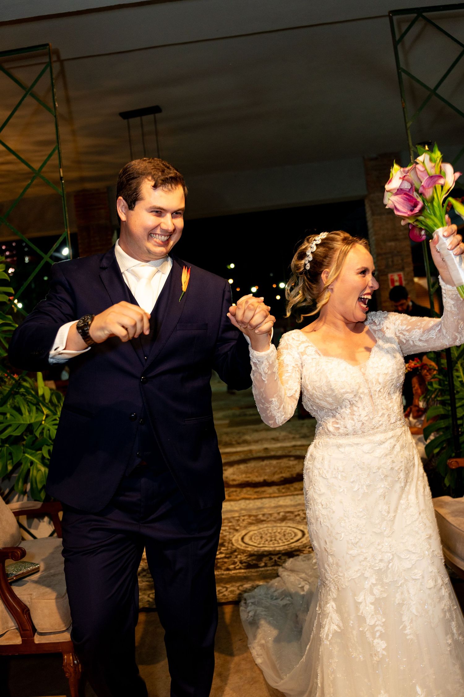 Joyful newlyweds celebrate after wedding ceremony, bride in lace gown holding pink bouquet, groom in navy suit.