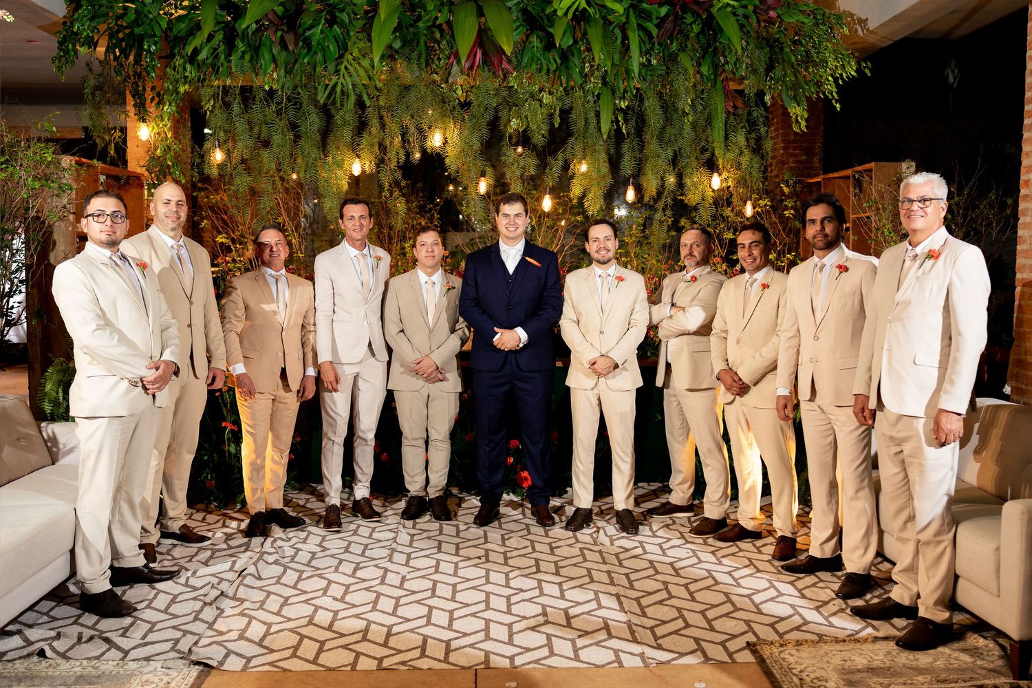 Group of men in beige and navy suits posing together at an elegant wedding venue with lush greenery.