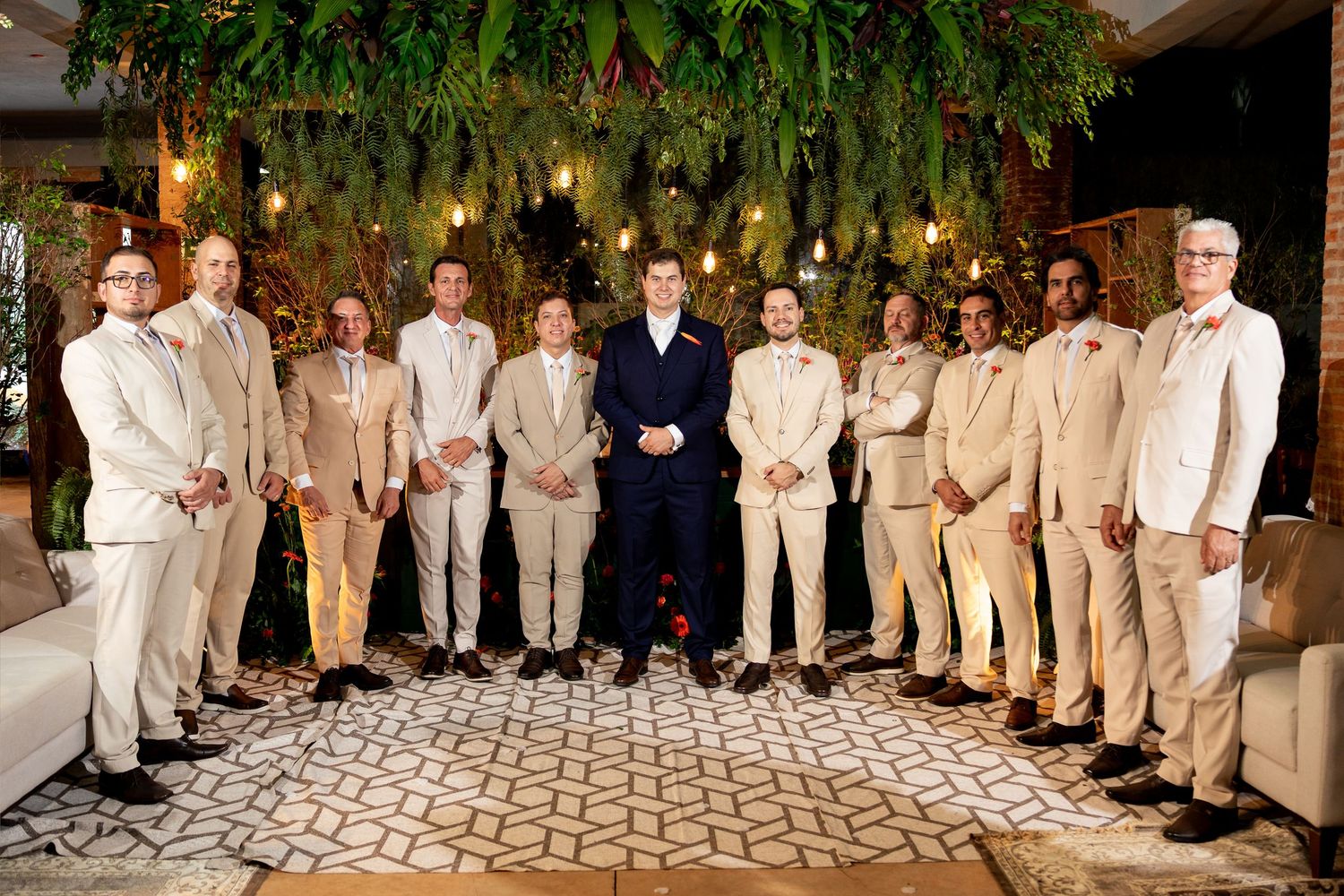 Groom in navy suit stands with groomsmen in tan suits at elegant wedding venue with lush greenery backdrop.