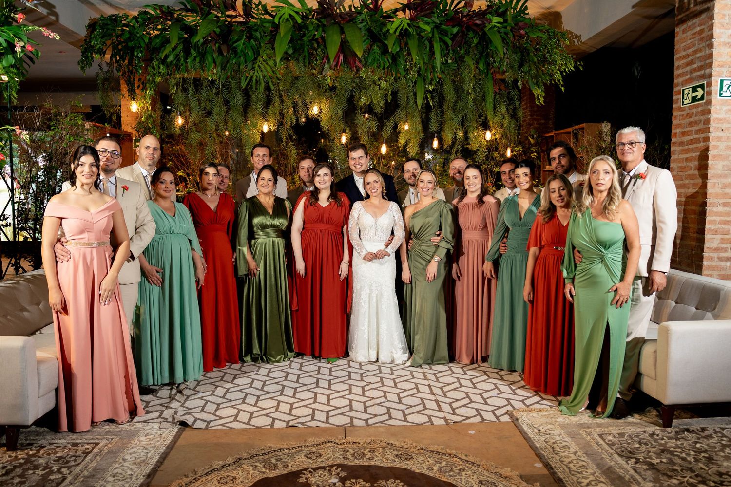 Wedding party poses in elegant venue with green and red bridesmaid gowns surrounding bride in white dress.