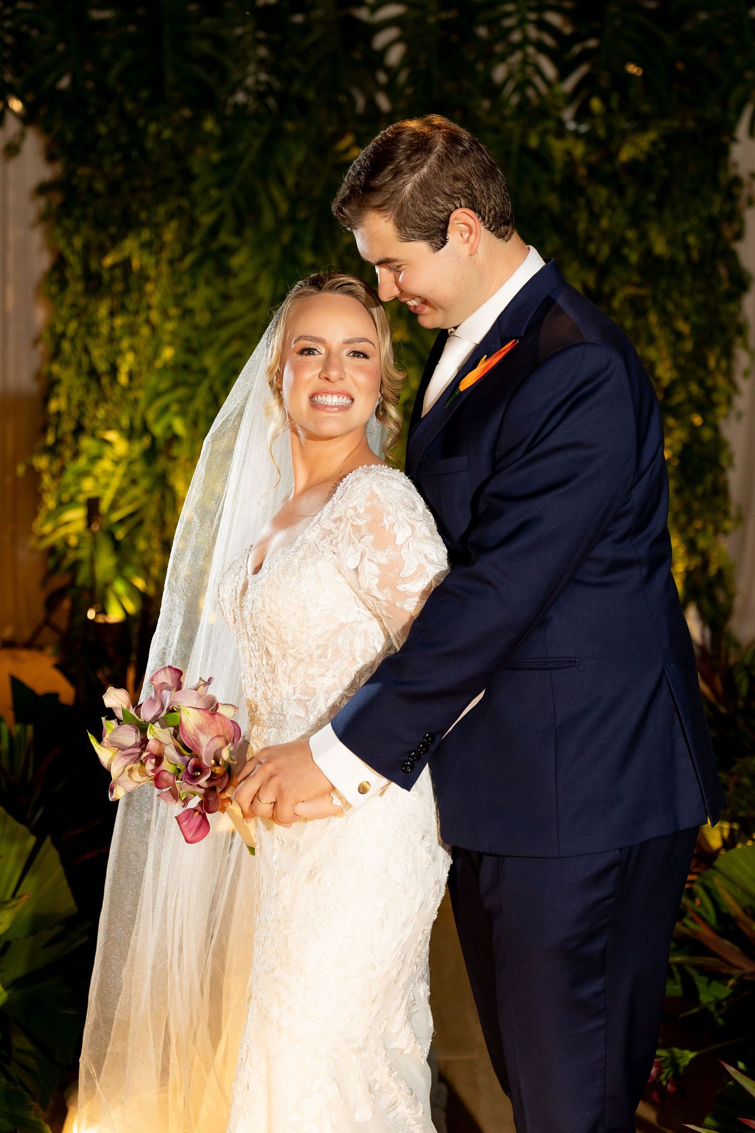 Smiling bride in lace gown with veil holds bouquet as groom in navy suit leans in tenderly, surrounded by lush greenery.