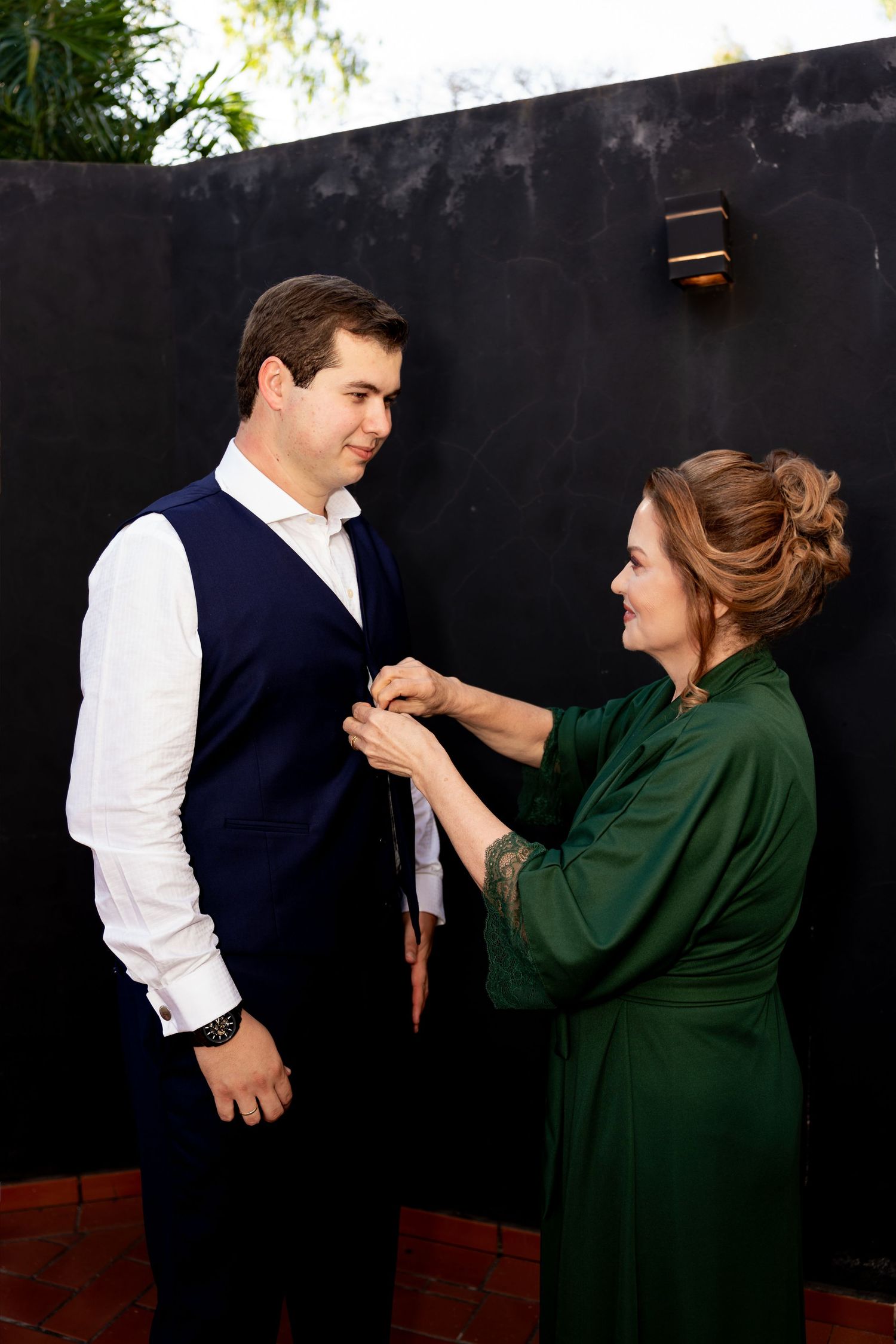 A woman in a green dress adjusts a mans tie as he stands in a navy vest and white shirt against a dark wall.