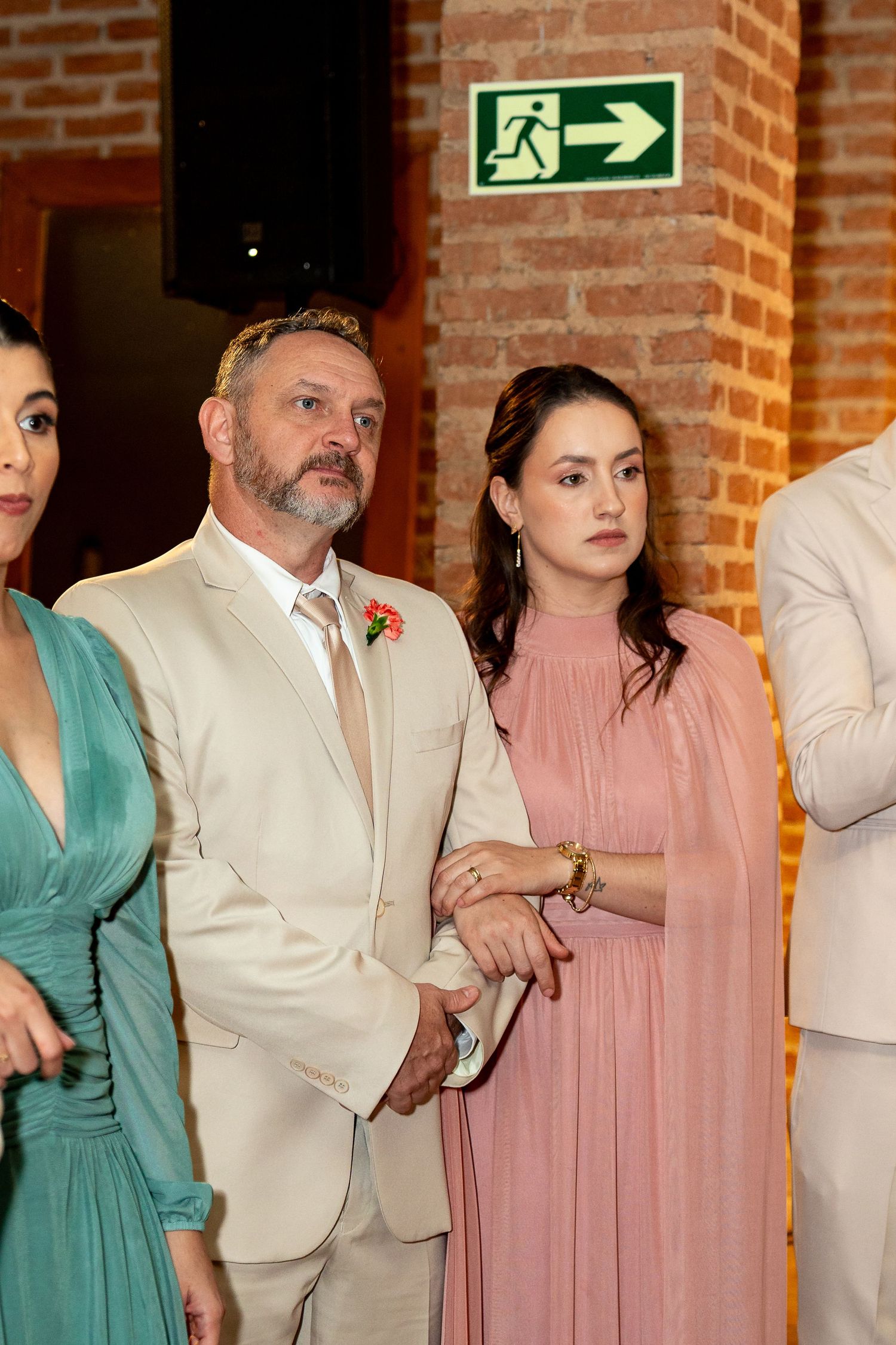 Elegantly dressed guests at a wedding, man in beige suit with boutonniere, women in teal and pink gowns.