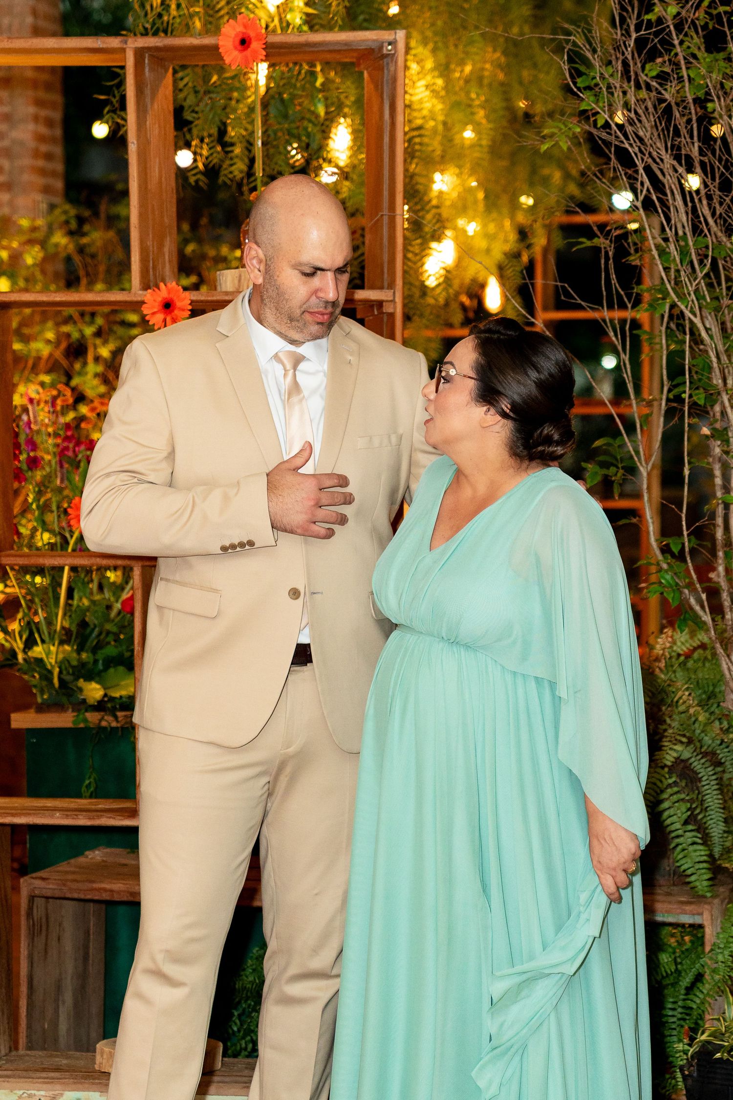 Couple in formal attire, man in beige suit and woman in teal gown, sharing a moment at an elegant event.