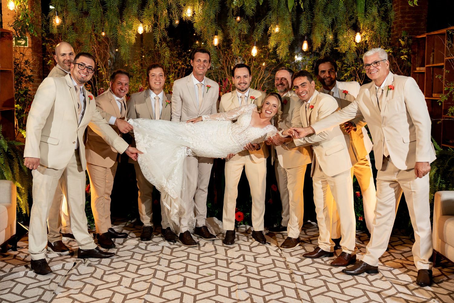 Groom and groomsmen in ivory suits lifting the bride in a white wedding dress outdoors at a festive venue.