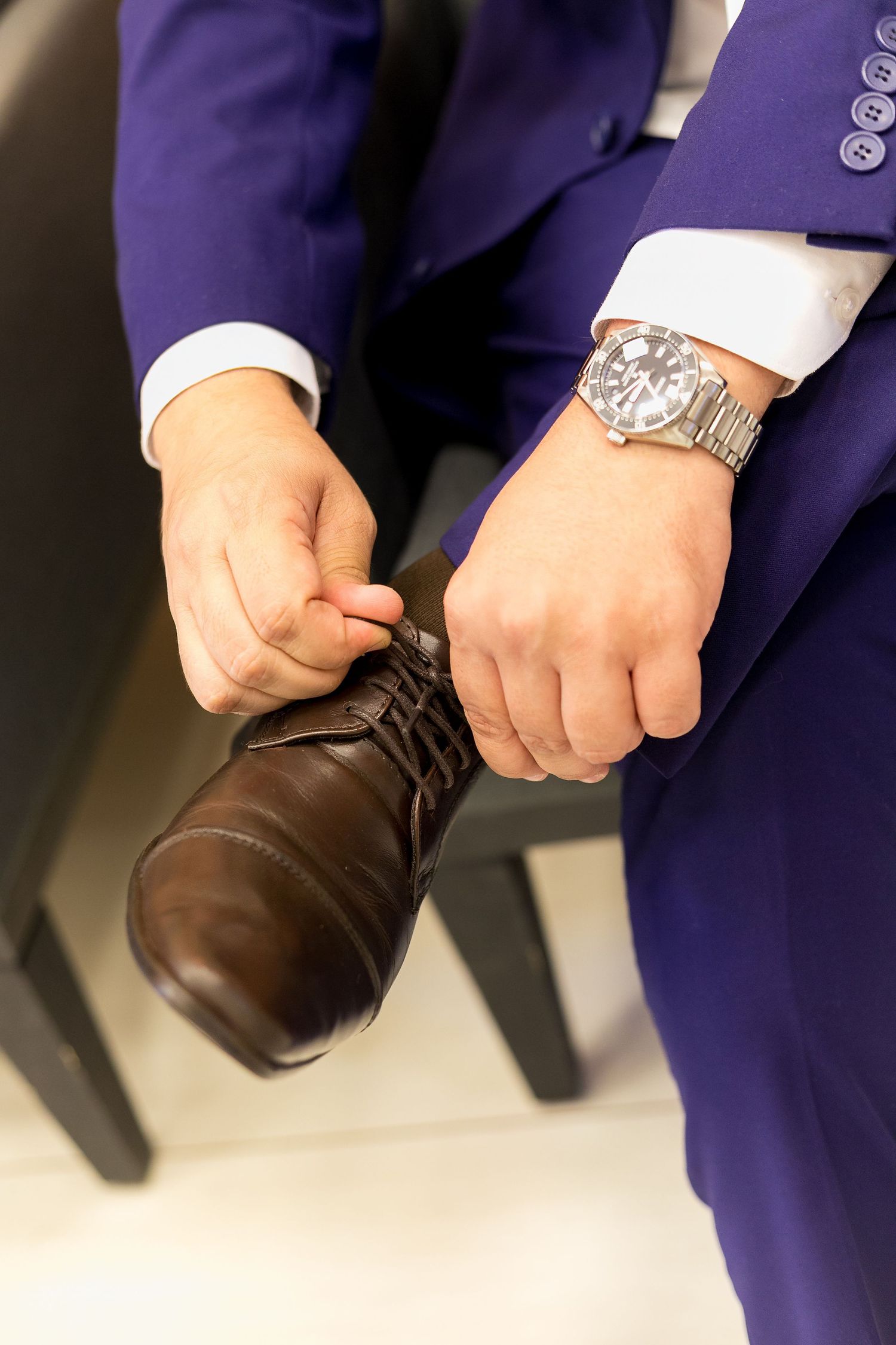 Man in blue suit and silver watch putting on brown leather dress shoe while seated.