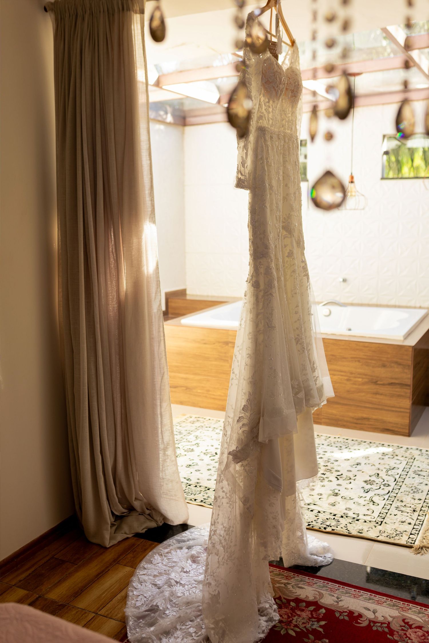 Elegant white lace wedding dress hanging on a wooden hanger in a sunlit bridal suite with hardwood floors.