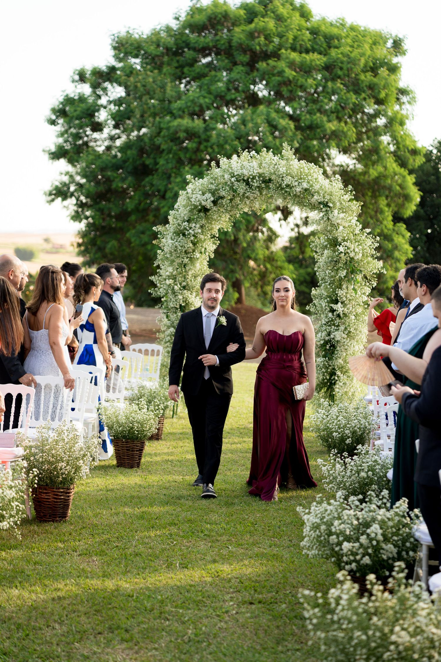 A couple walks down an outdoor wedding aisle under a floral arch, surrounded by guests on a lush green lawn.