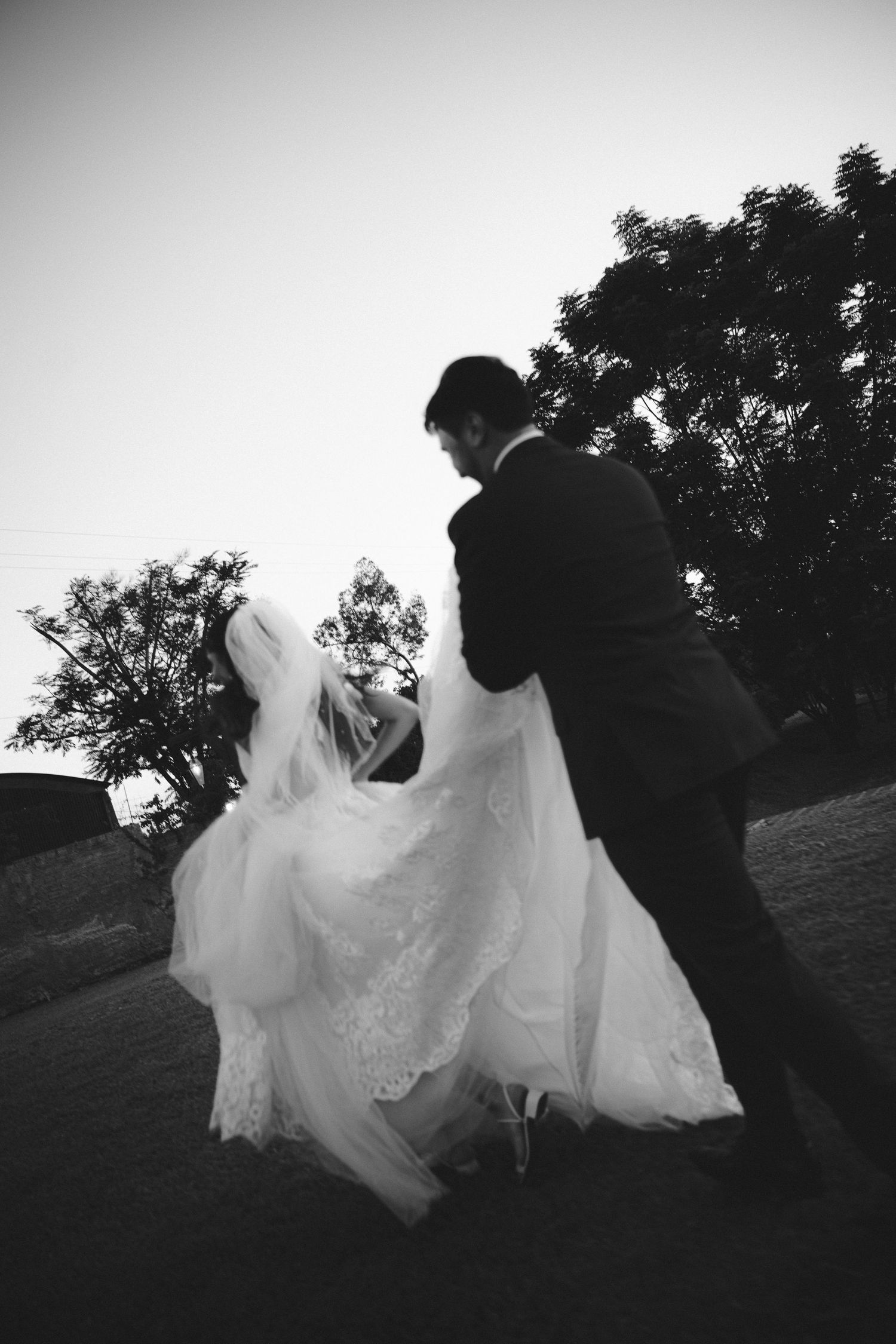 Black and white photo of a bride and groom dancing outdoors at dusk, with trees silhouetted against the sky.