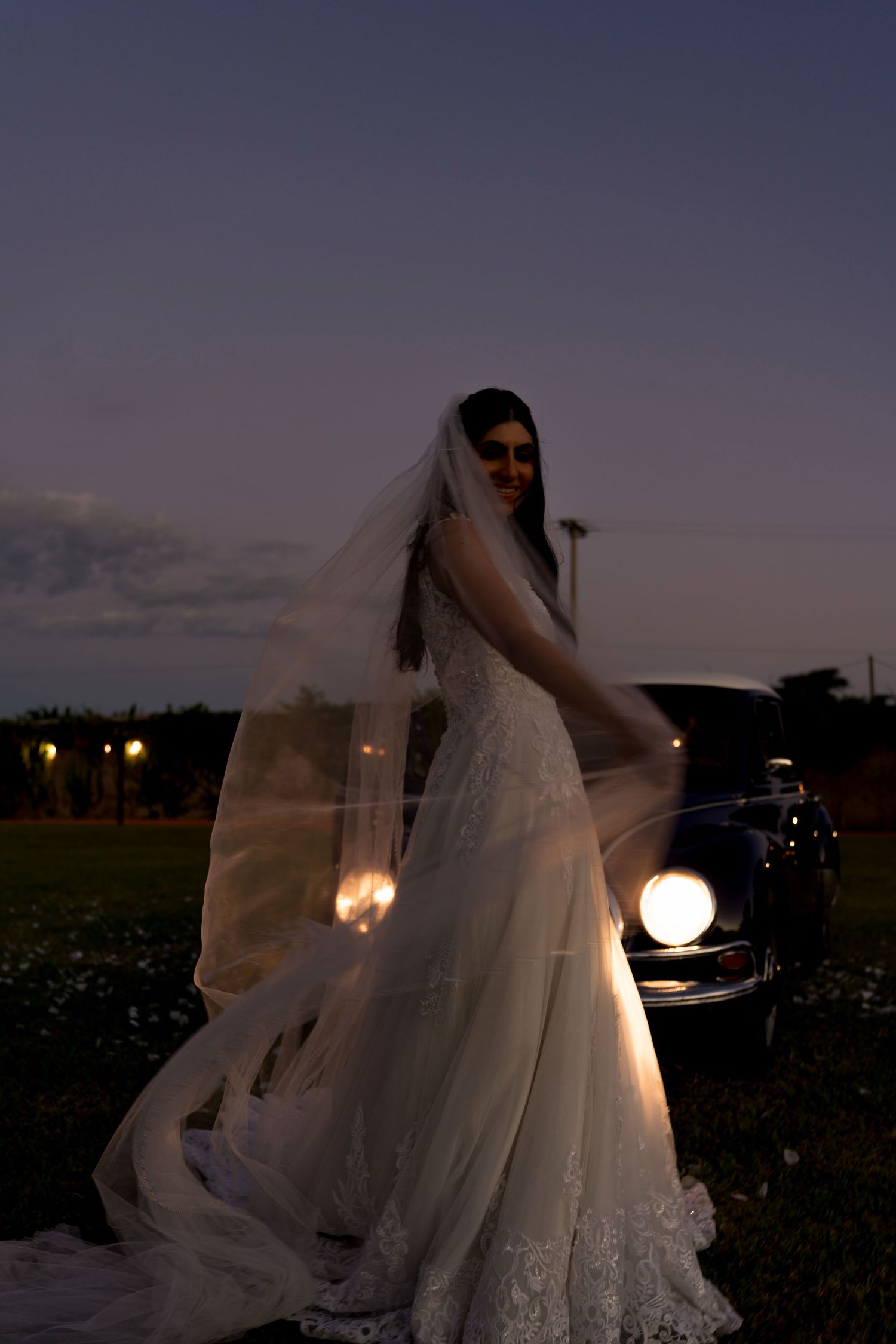 Bride in white lace gown and veil poses outdoors at dusk near vintage car with glowing headlights.