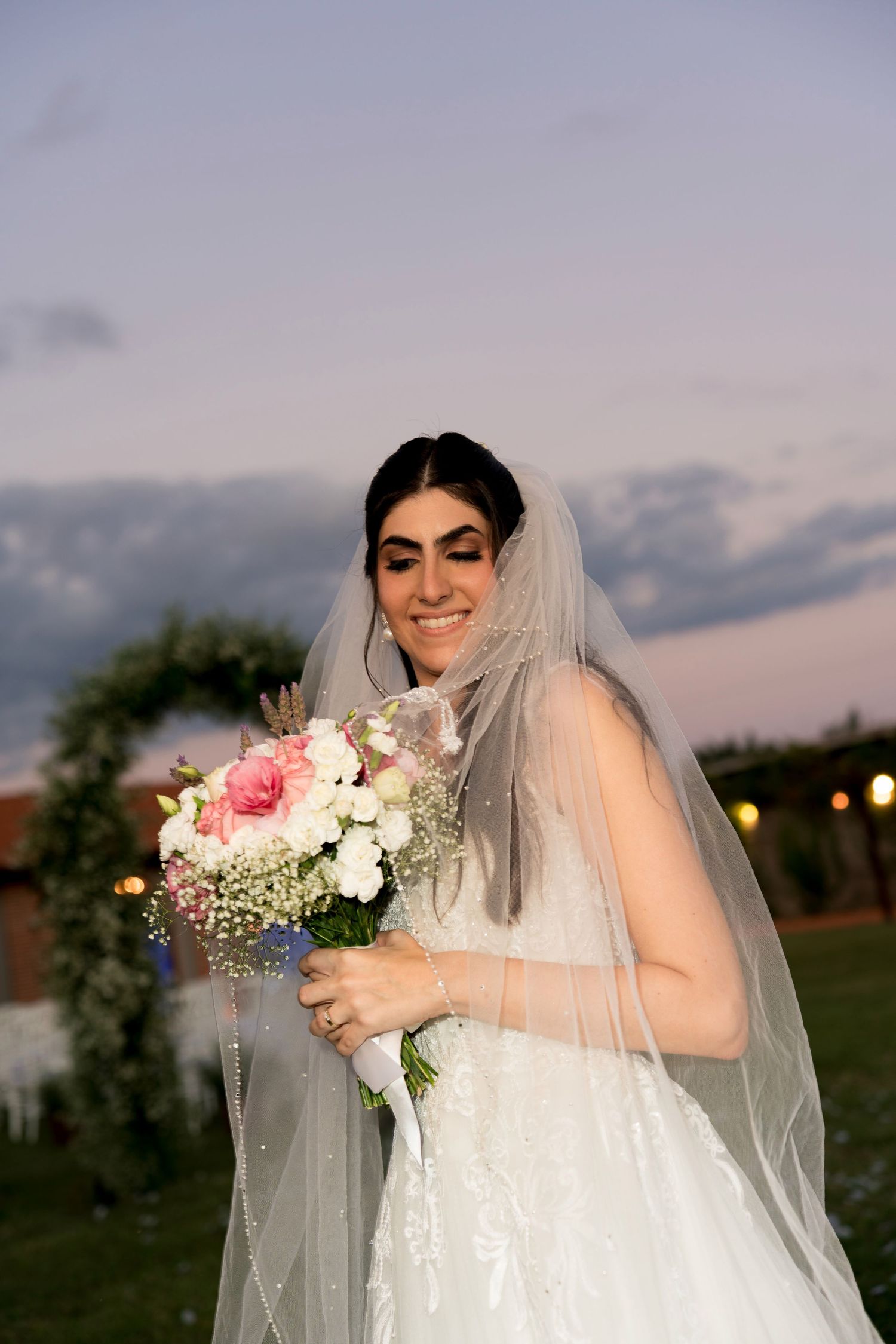 Smiling bride in white gown and veil holds pink and white bouquet outdoors at dusk.