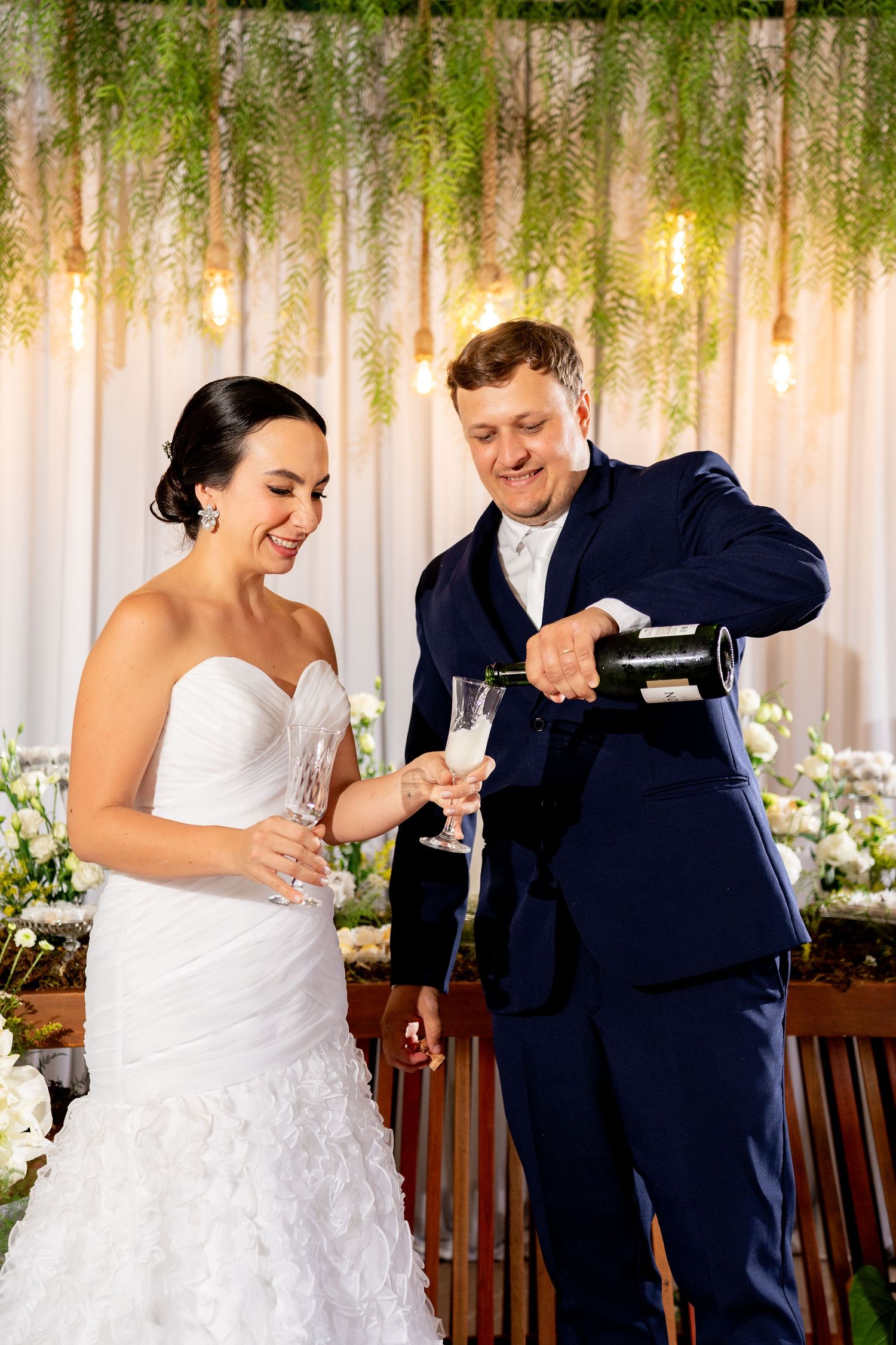 Bride and groom toasting with champagne glasses at wedding reception with floral backdrop.