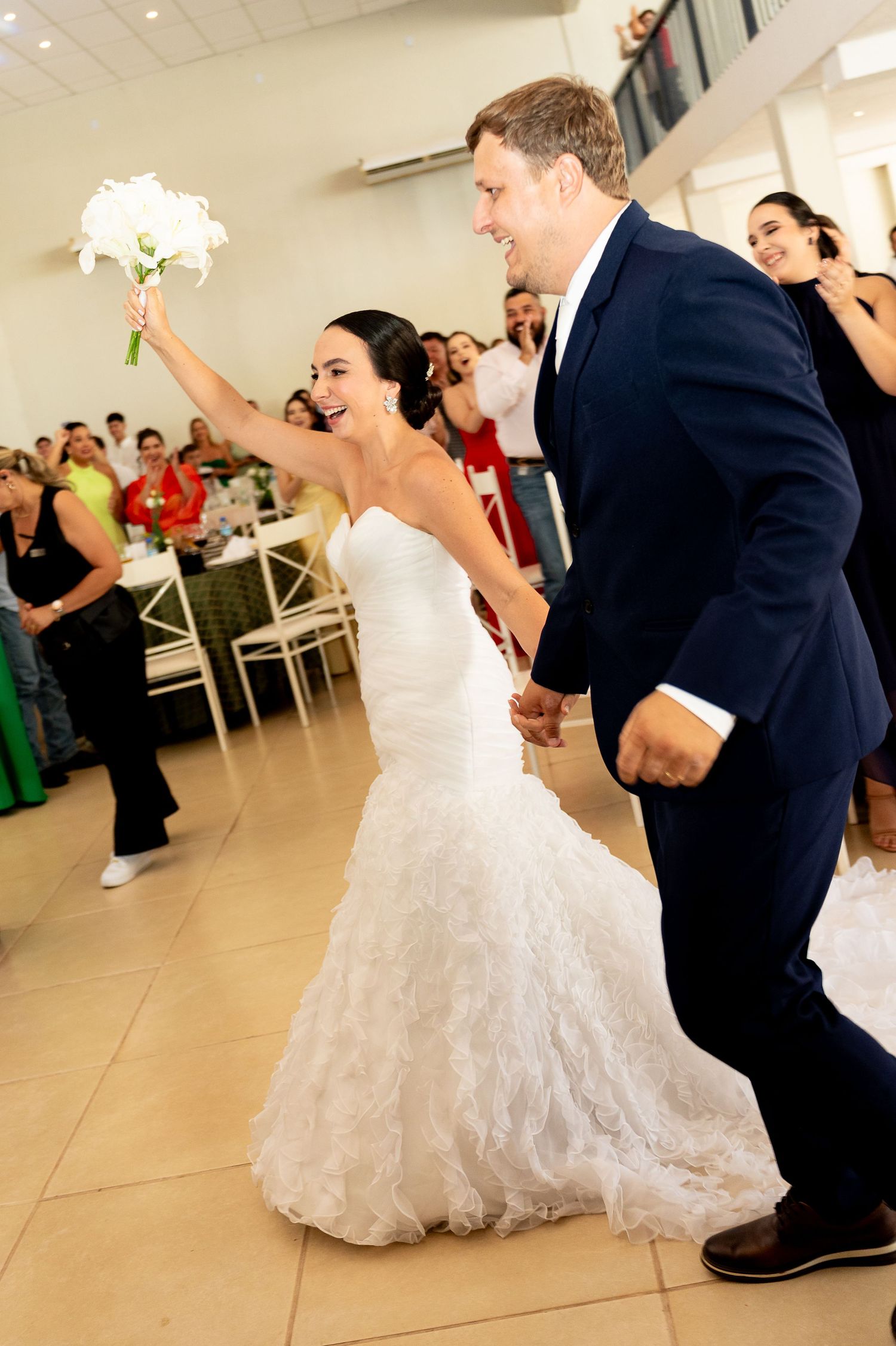 Joyful bride in white mermaid gown raises bouquet while groom in navy suit leads her onto wedding reception dance floor.
